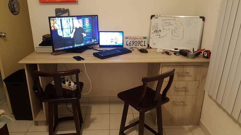 A cluttered desk with two chairs, one facing a desktop monitor showing a person on a stage, a laptop, a keyboard, and a mouse. The whiteboard has various sketches and notes. There are tools, a tape measure, and a license plate on the desk.
