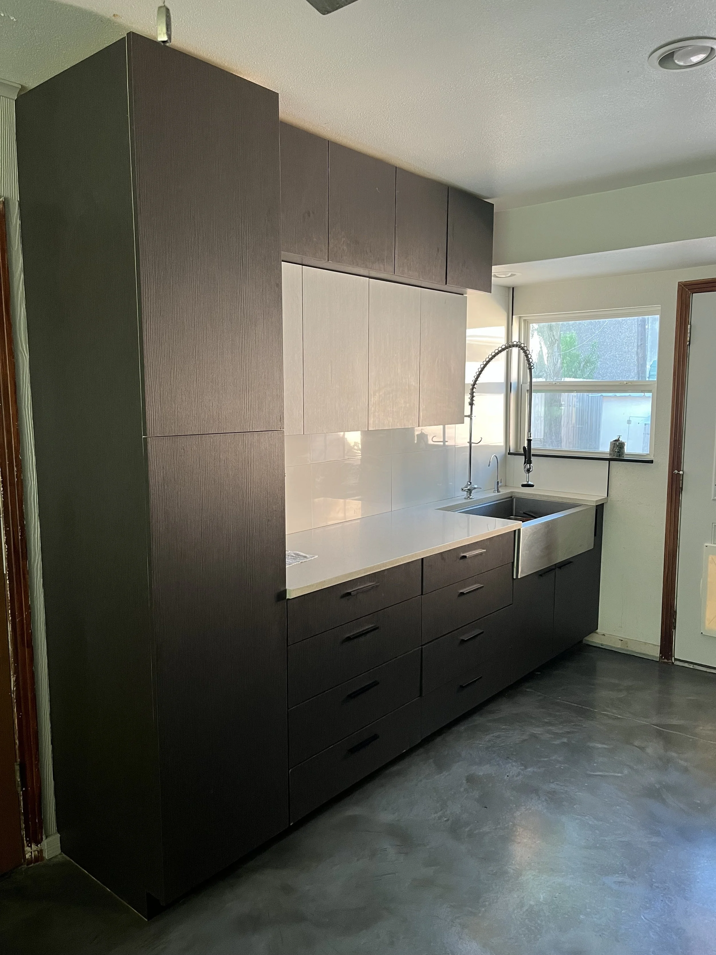 Modern kitchen with dark cabinet units, white countertop, stainless steel sink, tall window with natural light, black faucet, and dark flooring.