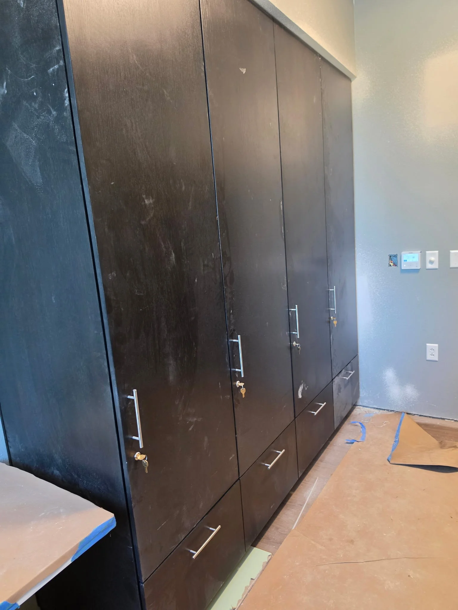 Black kitchen cabinets with silver handles, a beige countertop, and electrical outlets on the wall. The floor is brown with a protective covering on part of it.