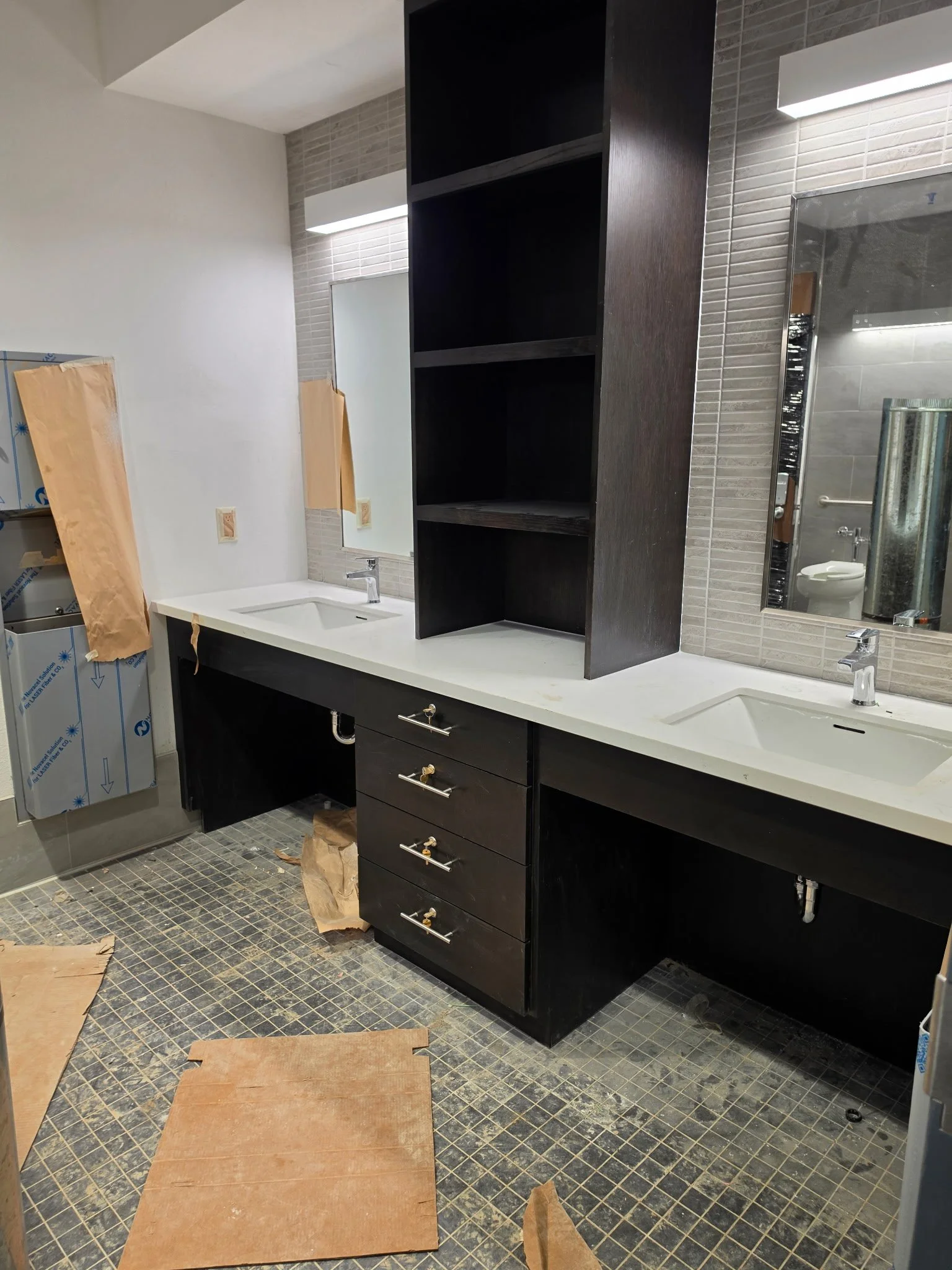 A bathroom under construction with black vanity cabinets, white countertops, double sinks, and unfinished flooring. There is a large mirror above the sinks, unfinished tiles, and construction materials on the floor.