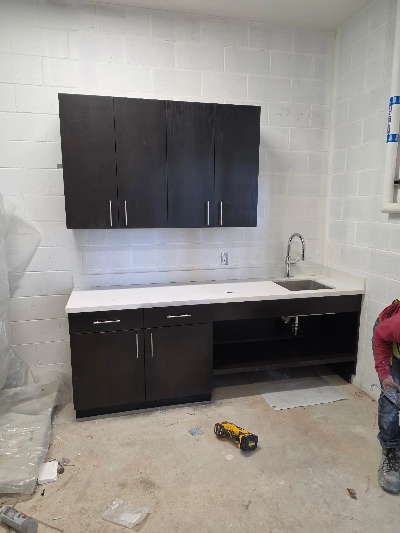 Kitchen construction site with black cabinets, a white countertop, a sink, and a worker's leg visible on the right side. Tools and debris are scattered on the unfinished floor.