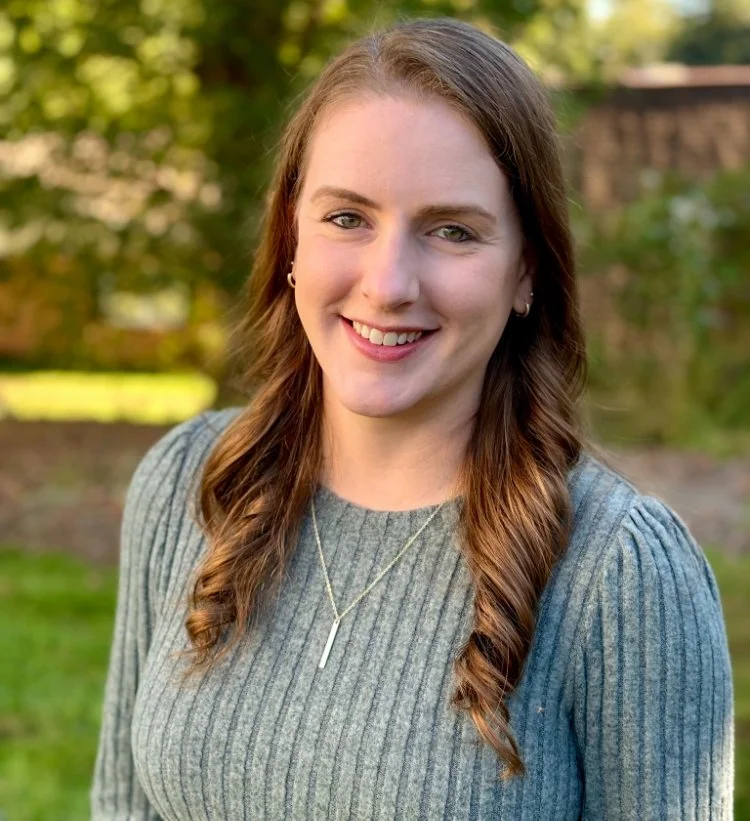 A young woman with long brown hair, smiling, wearing a gray knit sweater and a necklace, standing outdoors with blurred greenery in the background.