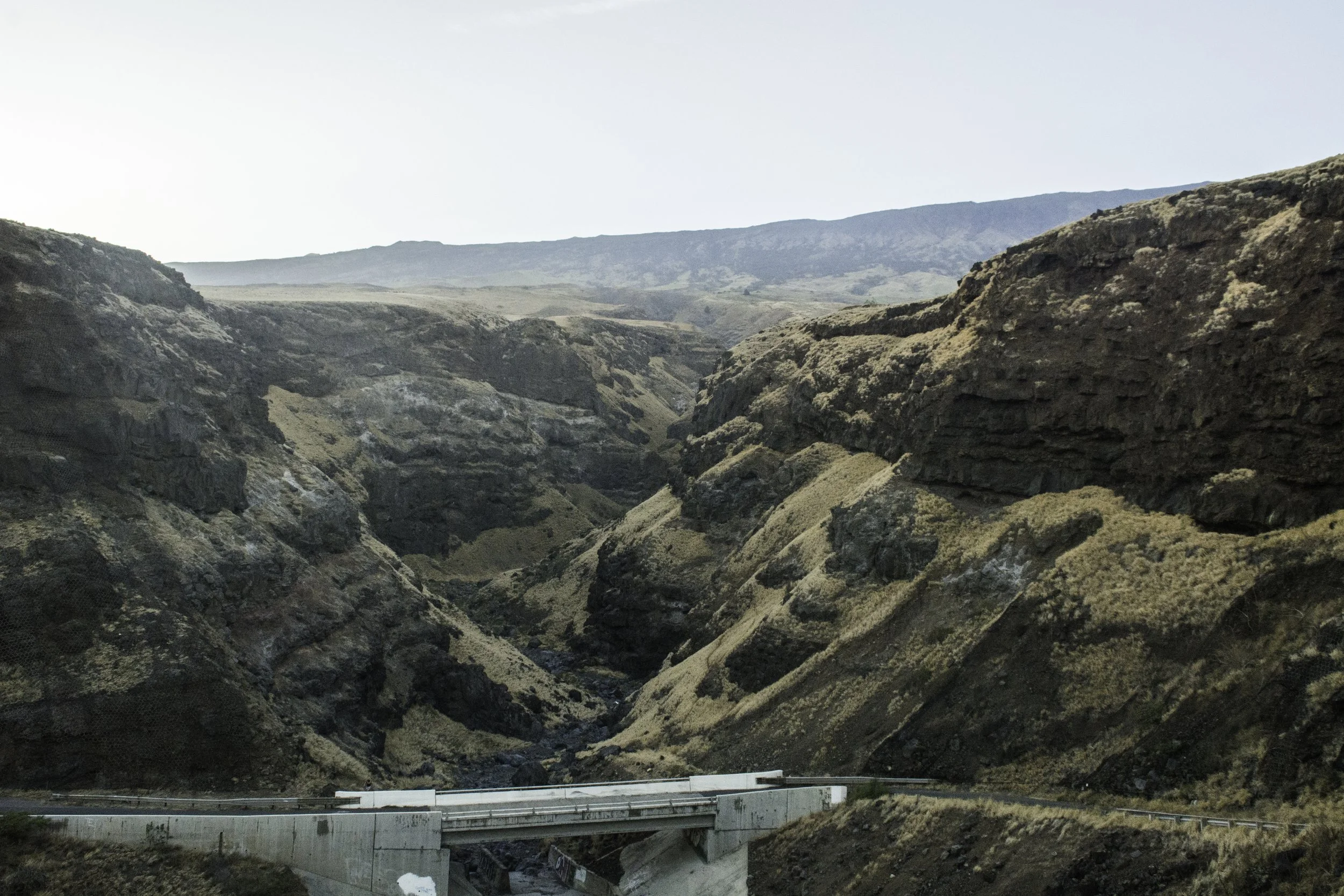 A mountainous landscape with rugged, steep cliffs and a road with a concrete barrier running through the valley at the bottom. Distant mountains are visible under a clear sky.