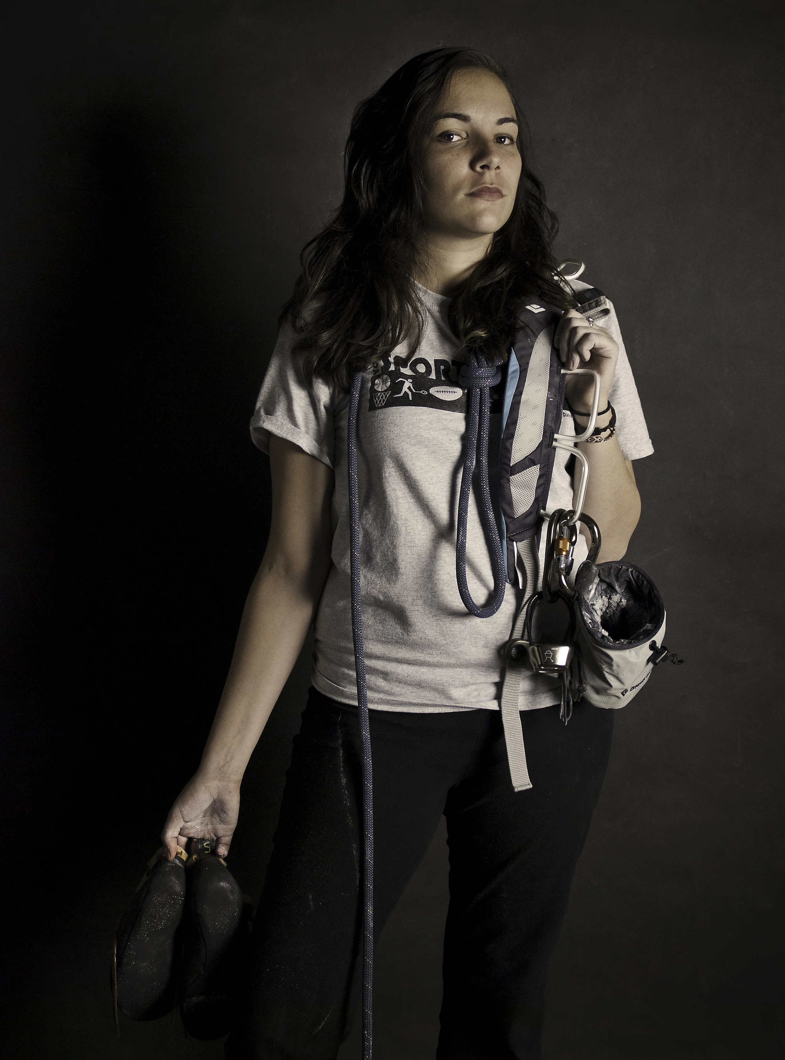 Young woman with dark wavy hair wearing a gray t-shirt with sports graphics carrying a backpack, holding black shoes in her left hand, standing against a dark background.