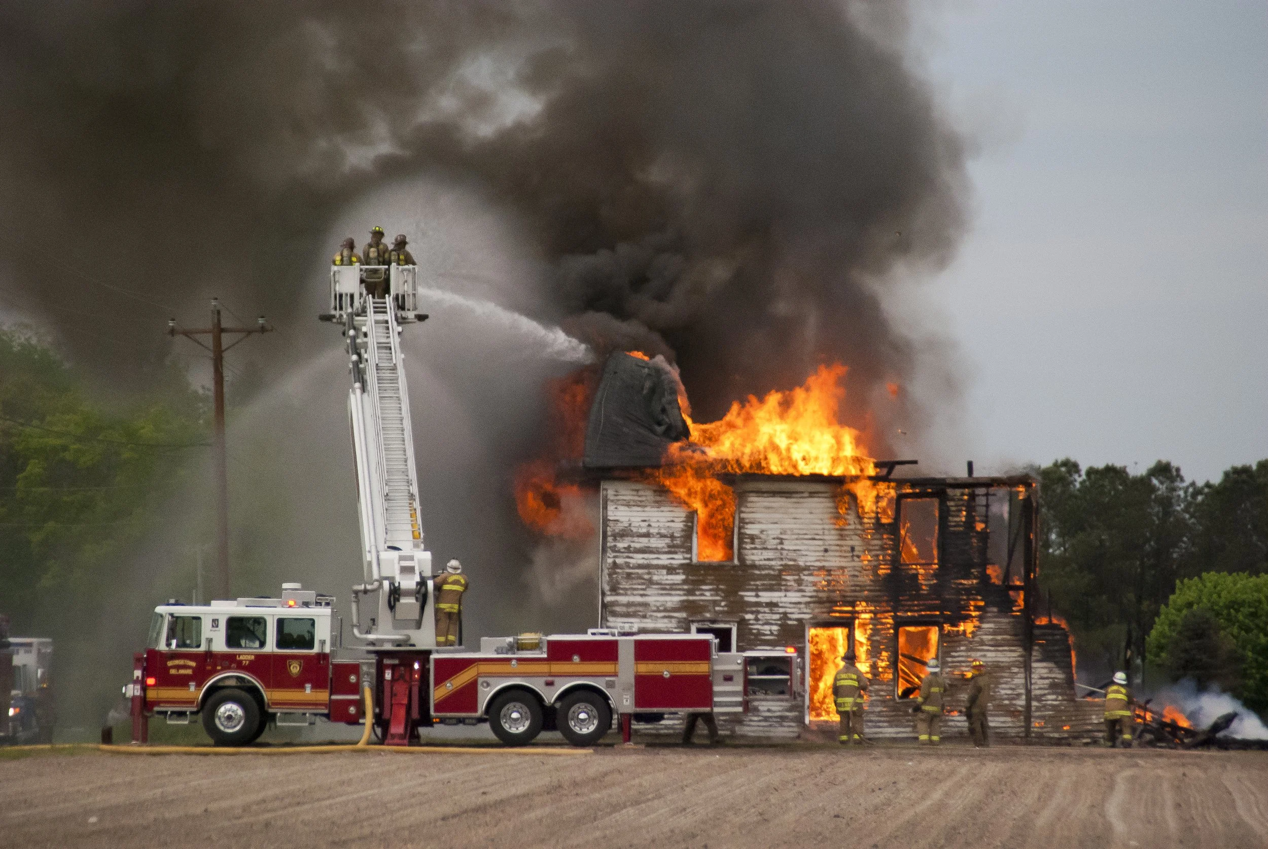 Firefighters are using an aerial ladder to spray water on a house that is engulfed in flames and thick black smoke.