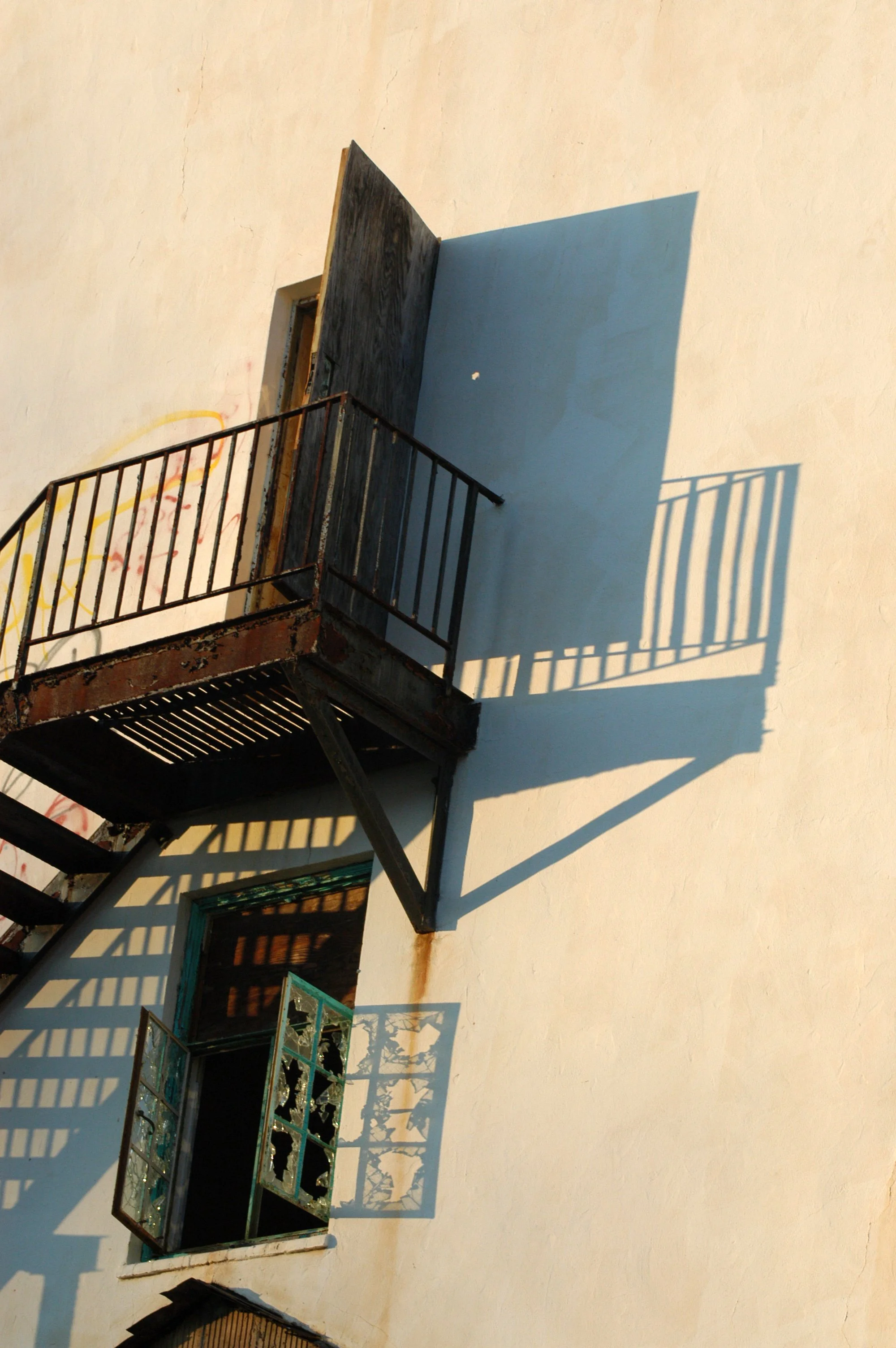 A white exterior wall with a black fire escape and a window with decorative glass, casting shadows on the wall.
