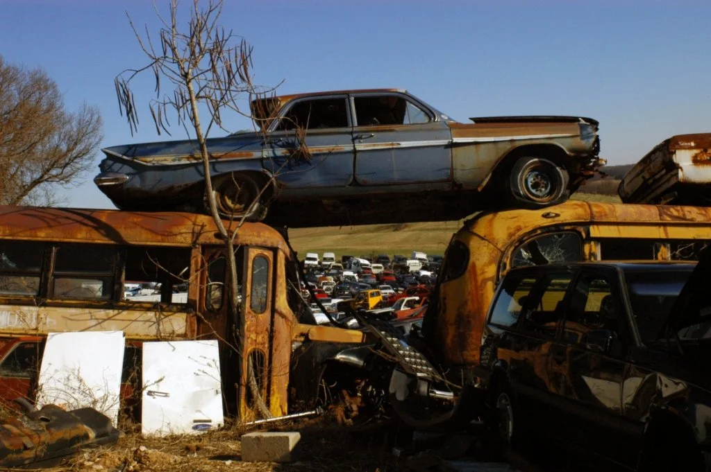 Stack of rusted, abandoned cars in a junkyard with a clear sky in the background.