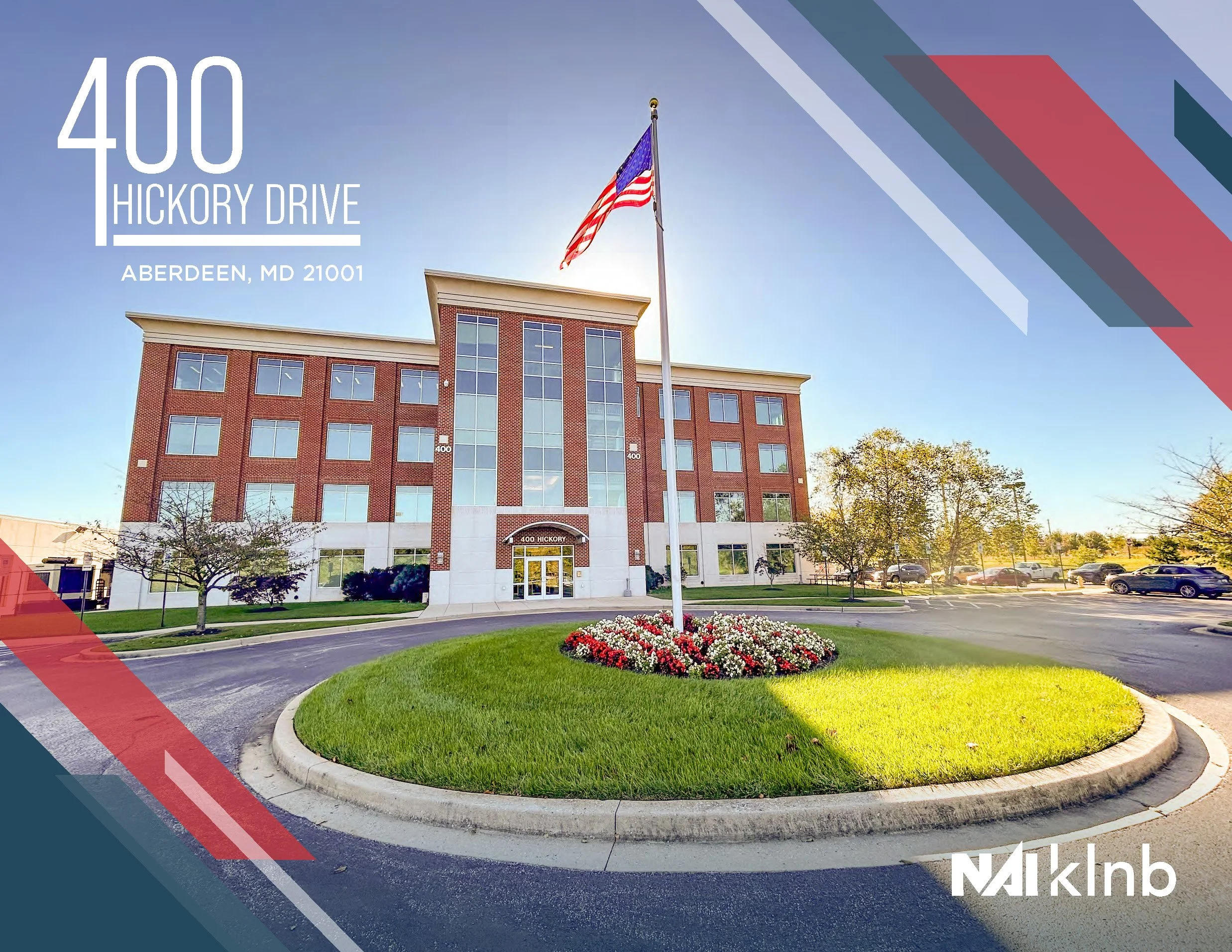 A multi-story brick office building with large windows, a flagpole with an American flag in front, and a landscaped circular flower bed with red, white, and pink flowers. The sky is clear and blue.
