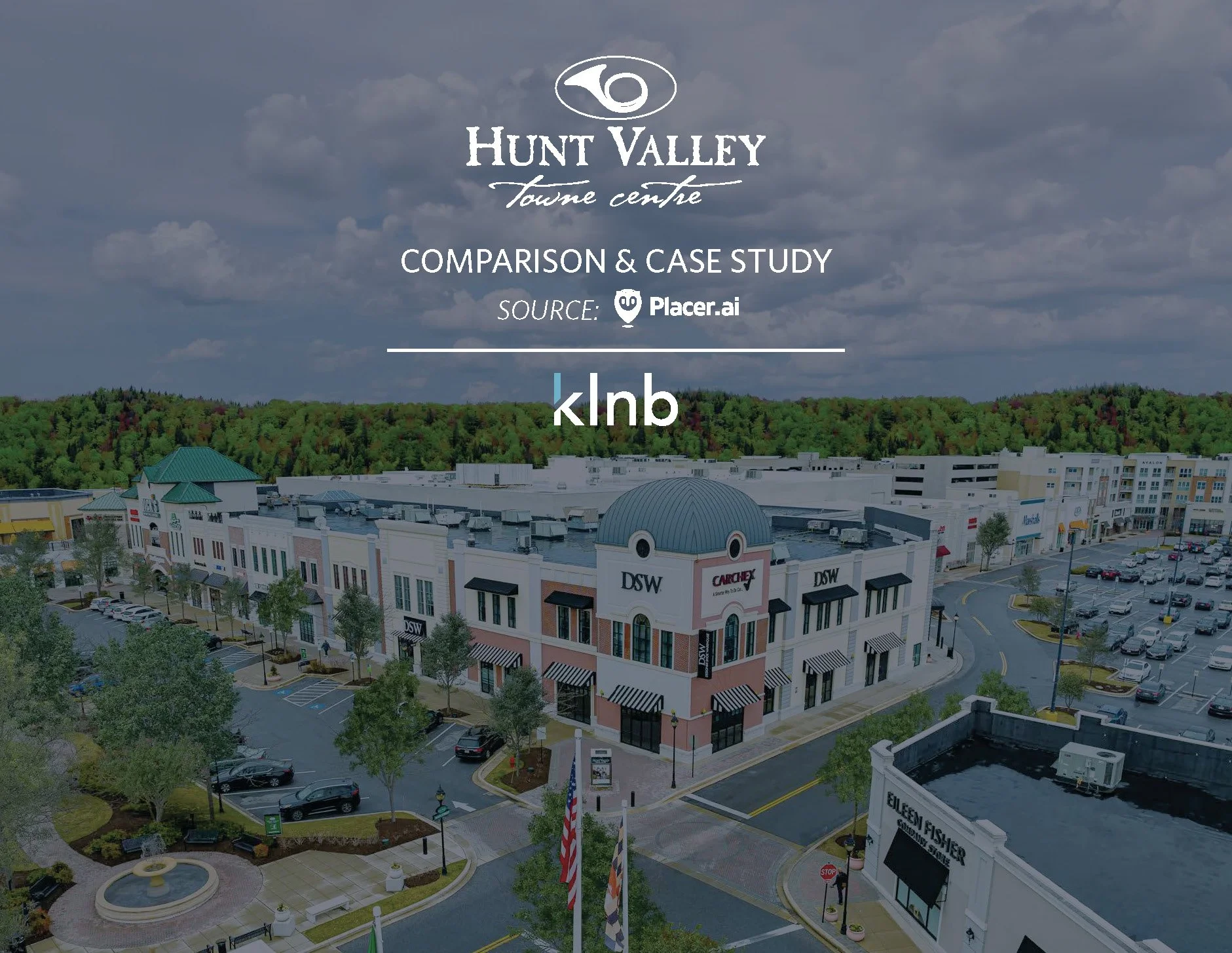 An aerial view of Hunt Valley Towne Centre shopping area with boats in a small pond, American flags, and parking lots filled with cars, surrounded by trees and commercial buildings.