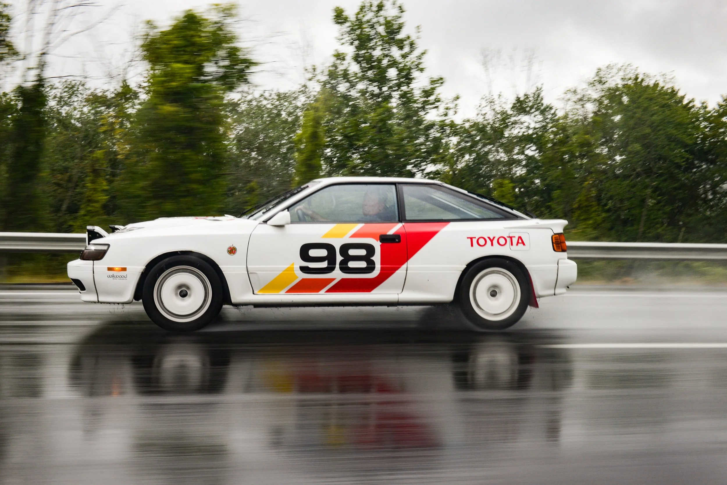 A white vintage Toyota race car with the number 98 and colorful racing stripes is driving on a wet road with a blurred background of trees.