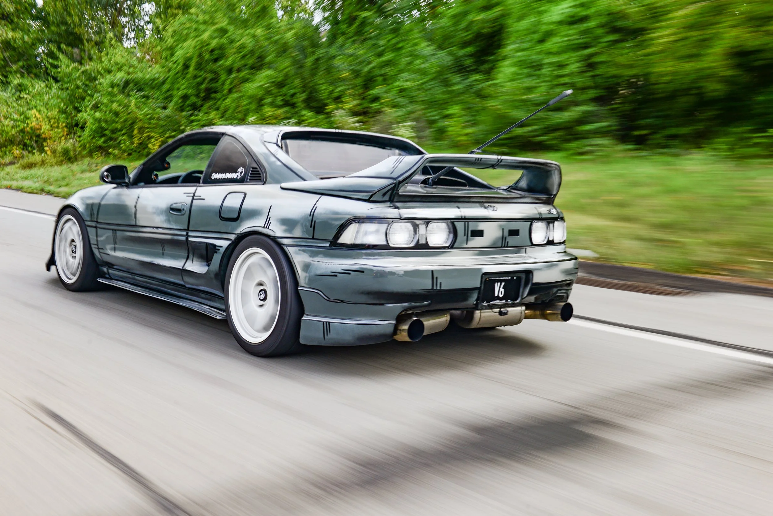 A silver sports car with a large rear spoiler driving on a highway with green trees in the background.