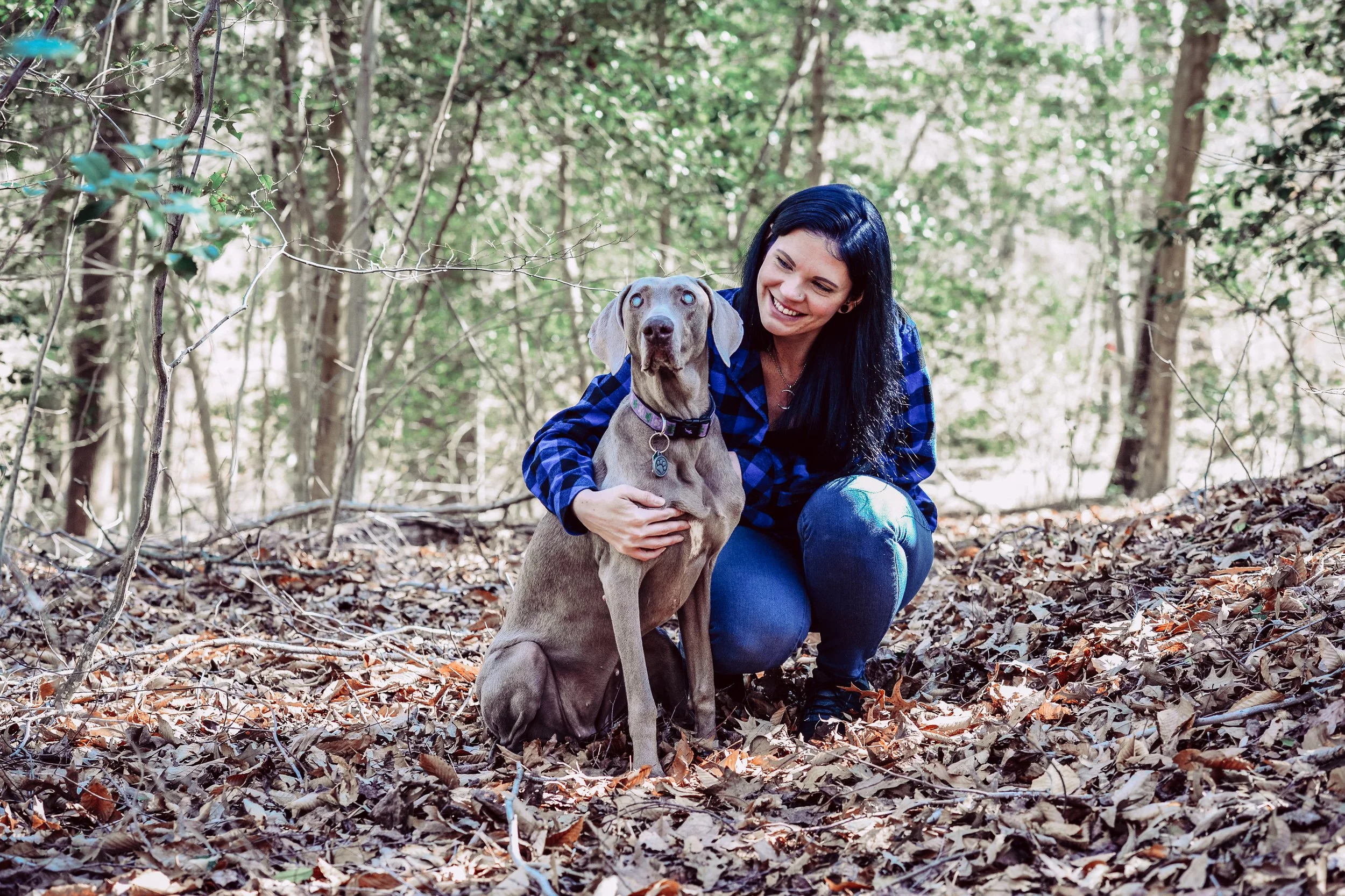 A woman with black hair smiling and crouching next to a large gray dog with blue eyes, in a wooded area with fallen leaves on the ground.