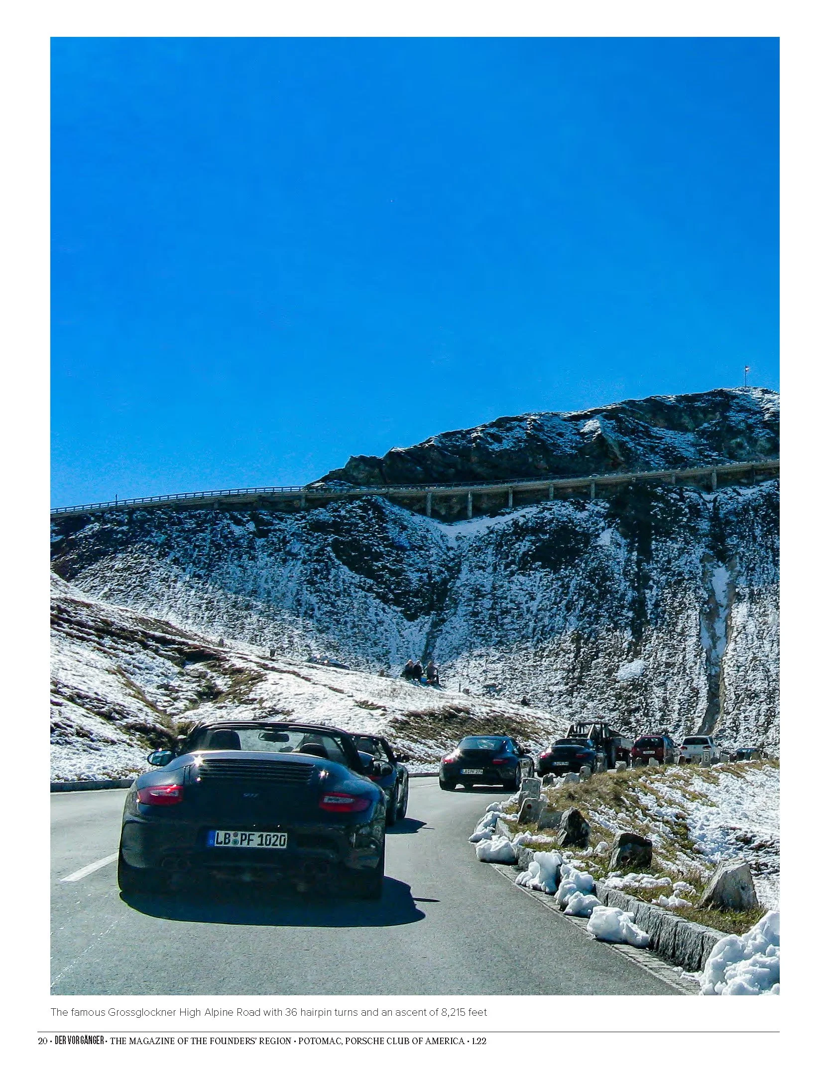 Cars parked along a road with snow on the ground, mountains in the background, and a clear blue sky.