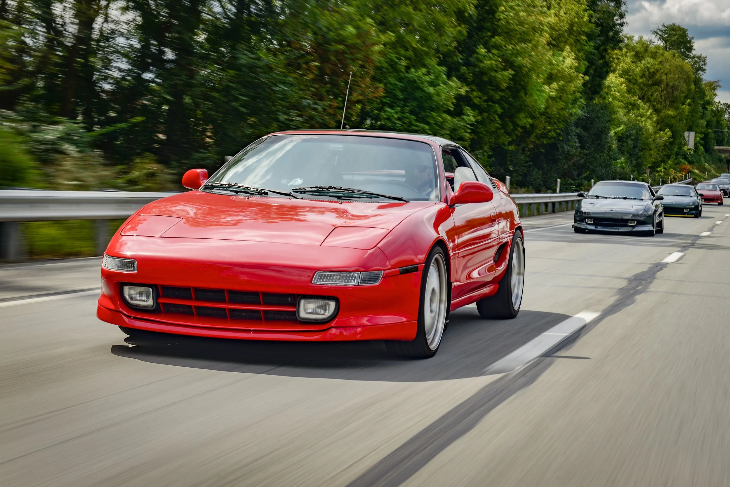 A red sports car driving on a highway with other cars behind it, surrounded by green trees and a cloudy sky.