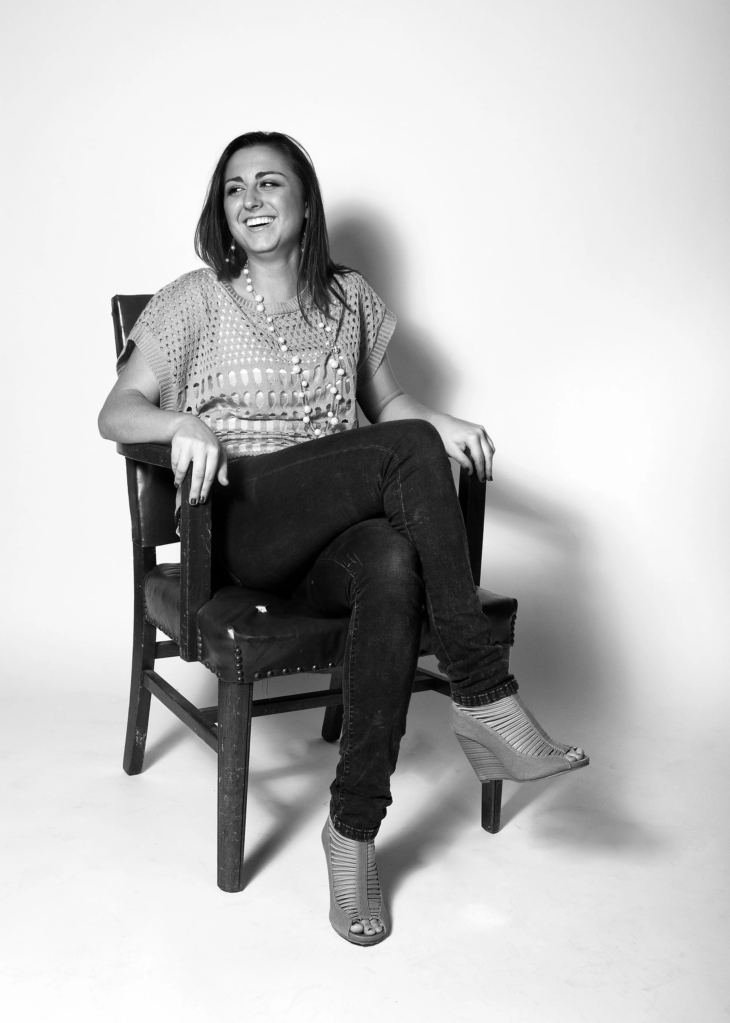 A woman with shoulder-length dark hair sitting on a wooden chair, smiling and looking to her left. She is wearing a light, open-weave top, dark jeans, and high-heeled sandals. The background is plain white.