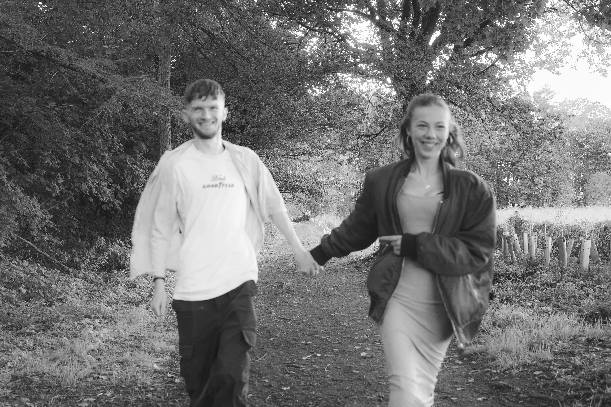 A black-and-white photo of a young man and woman holding hands and walking along a wooded trail. The man has short, curly hair and a beard, and the woman has long hair and is smiling. Trees and bushes surround the path.