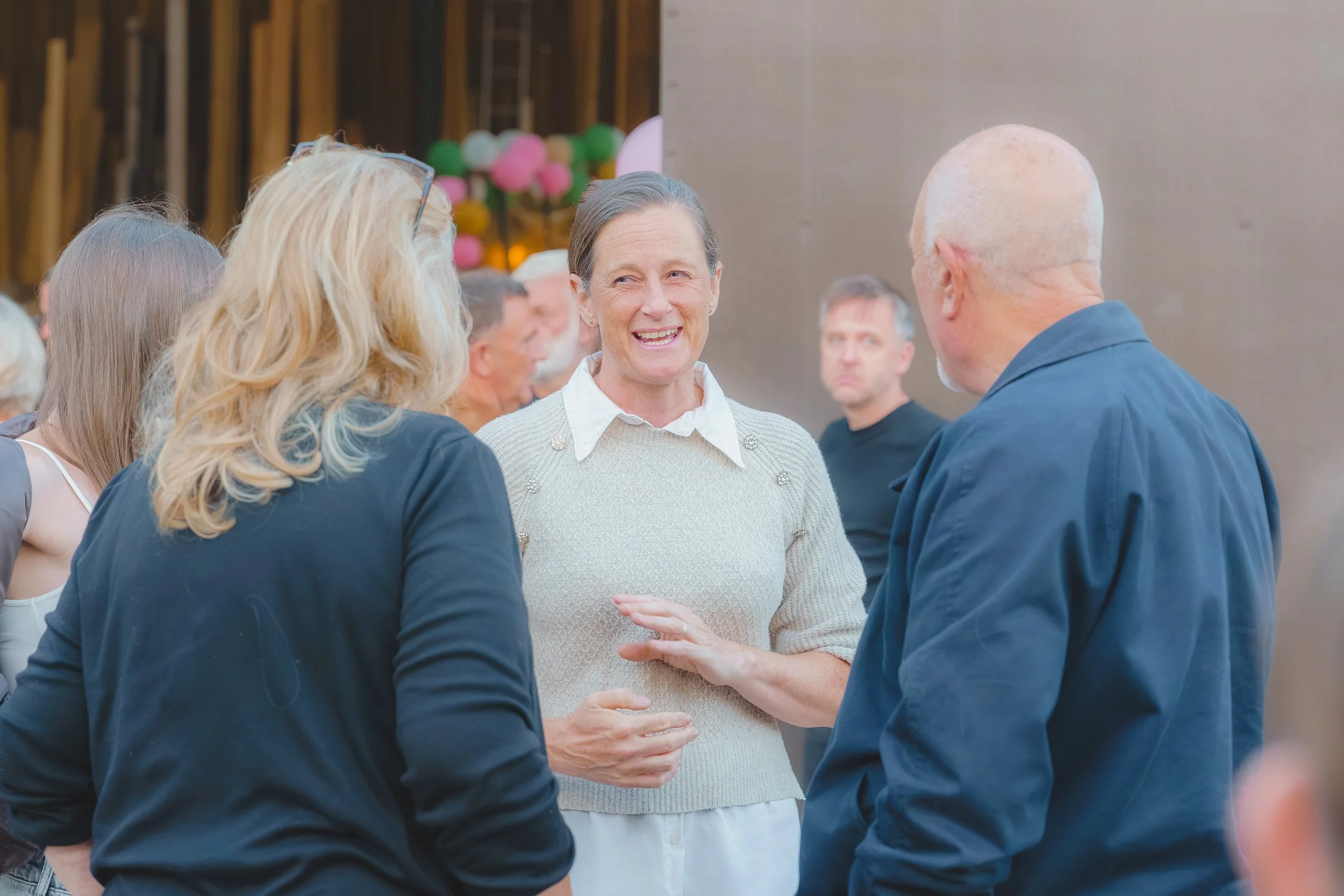 A group of people at a social gathering, with a woman in a white sweater smiling and talking to a man in a dark jacket, while others listen and engage in conversation in the background.