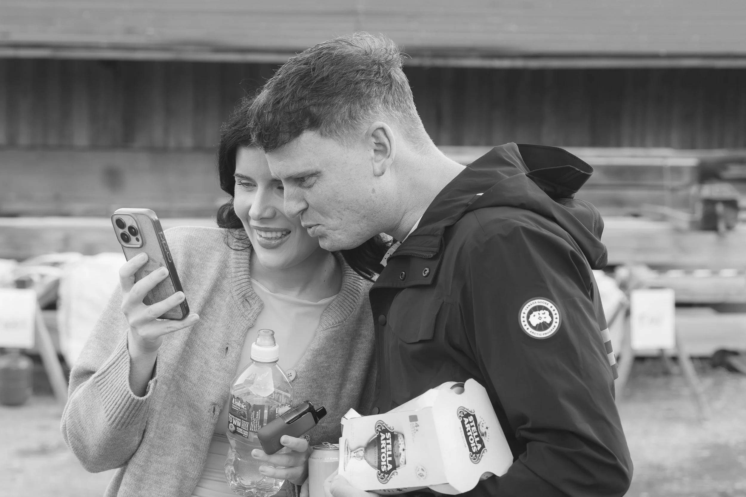 A woman and a man look at a phone together at an outdoor market, with food items in the man's hands, in black and white.