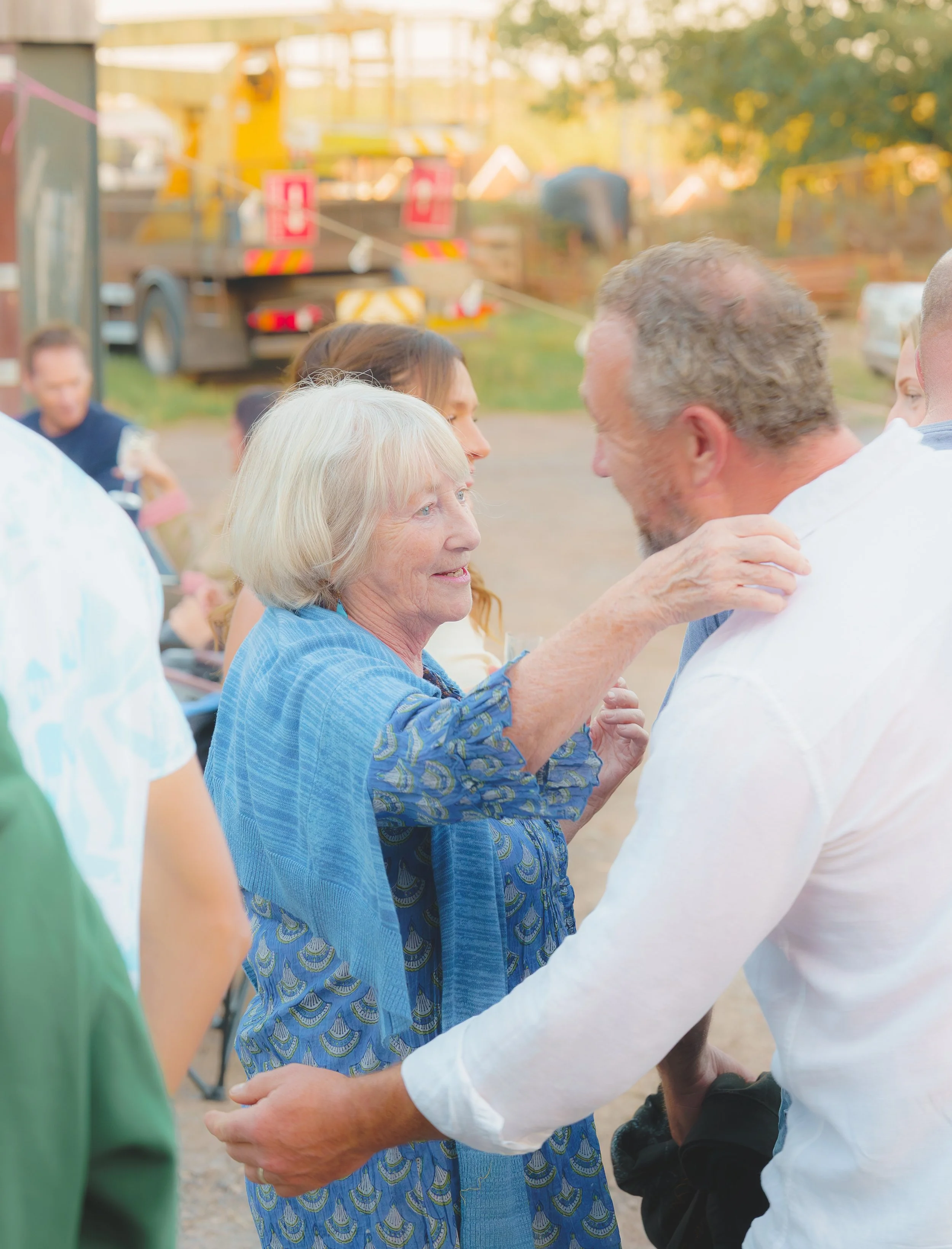 An elderly woman and a man are embracing and holding hands outdoors at a gathering. The background has a blurry play area with a yellow slide, and other people are present.