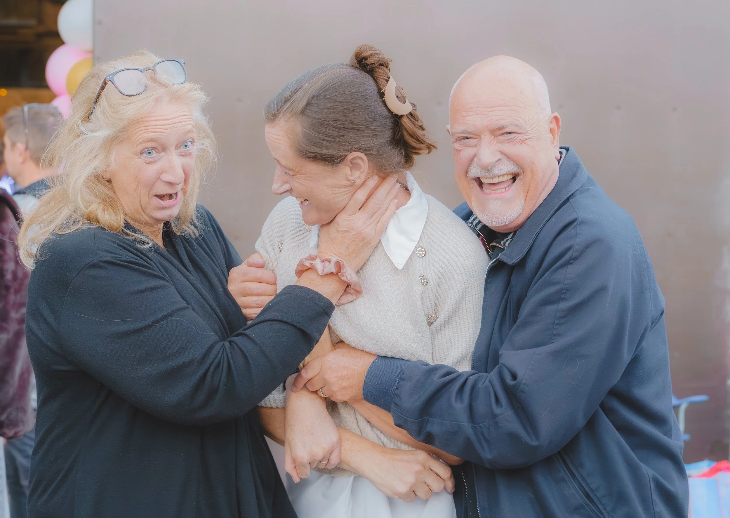 Three older adults, two women and one man, are laughing and hugging each other at what appears to be a celebration or gathering. They are smiling, and the atmosphere looks joyful.