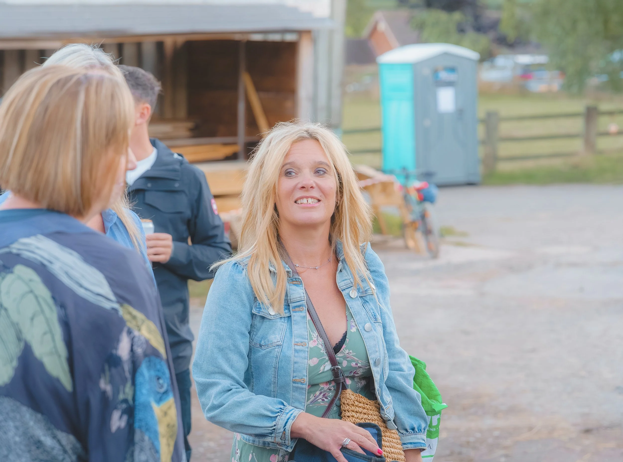 Three women and one man engaged in conversation outdoors, with trees, a wooden structure, and a portable toilet in the background. One woman has blonde hair, is wearing a denim jacket, and is smiling. The others are partially visible.