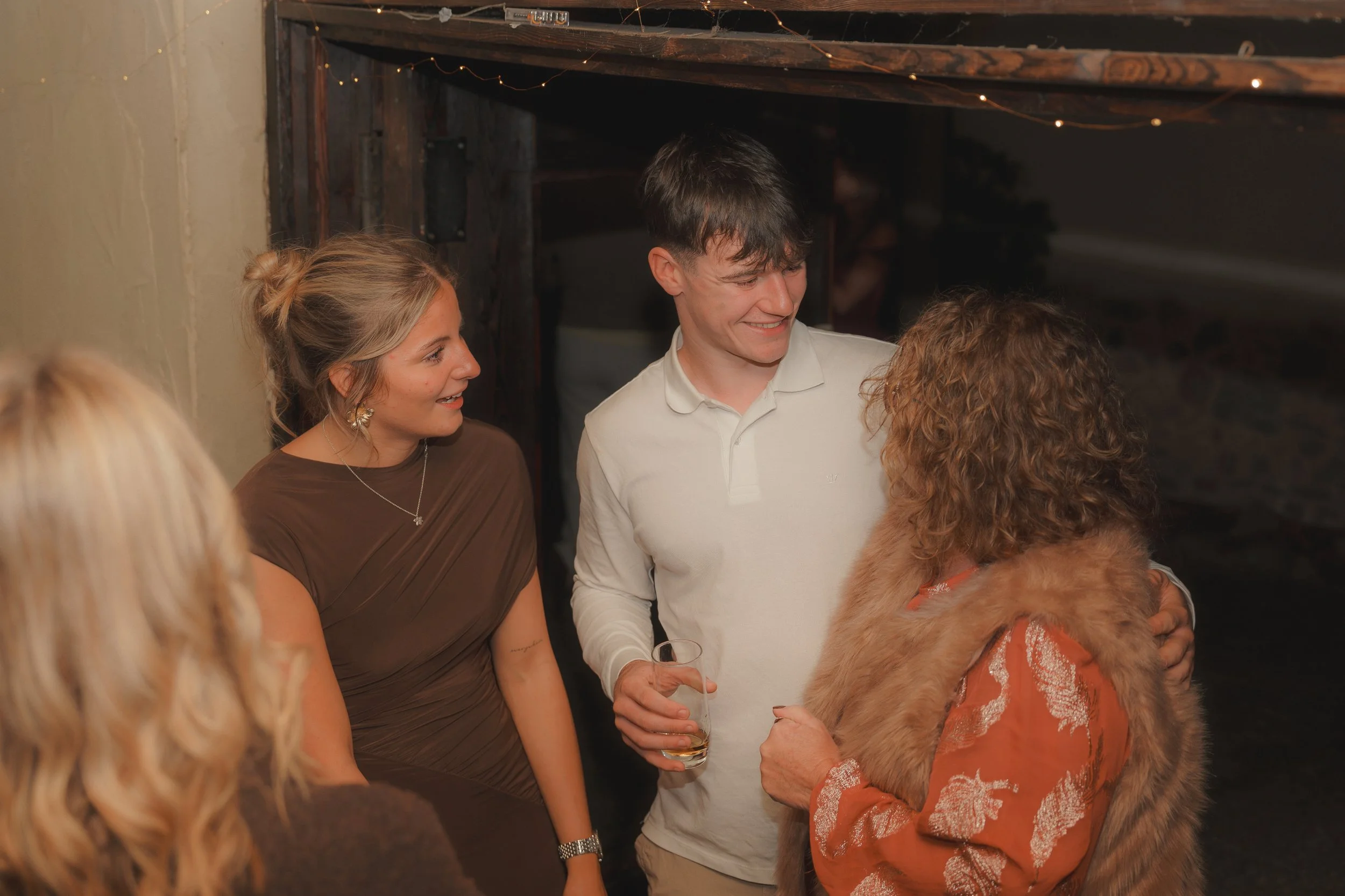 Group of people socializing at an indoor gathering, with a woman in a brown dress talking to a man in a white shirt holding a drink, and a woman with curly hair and a furry coat in the foreground.