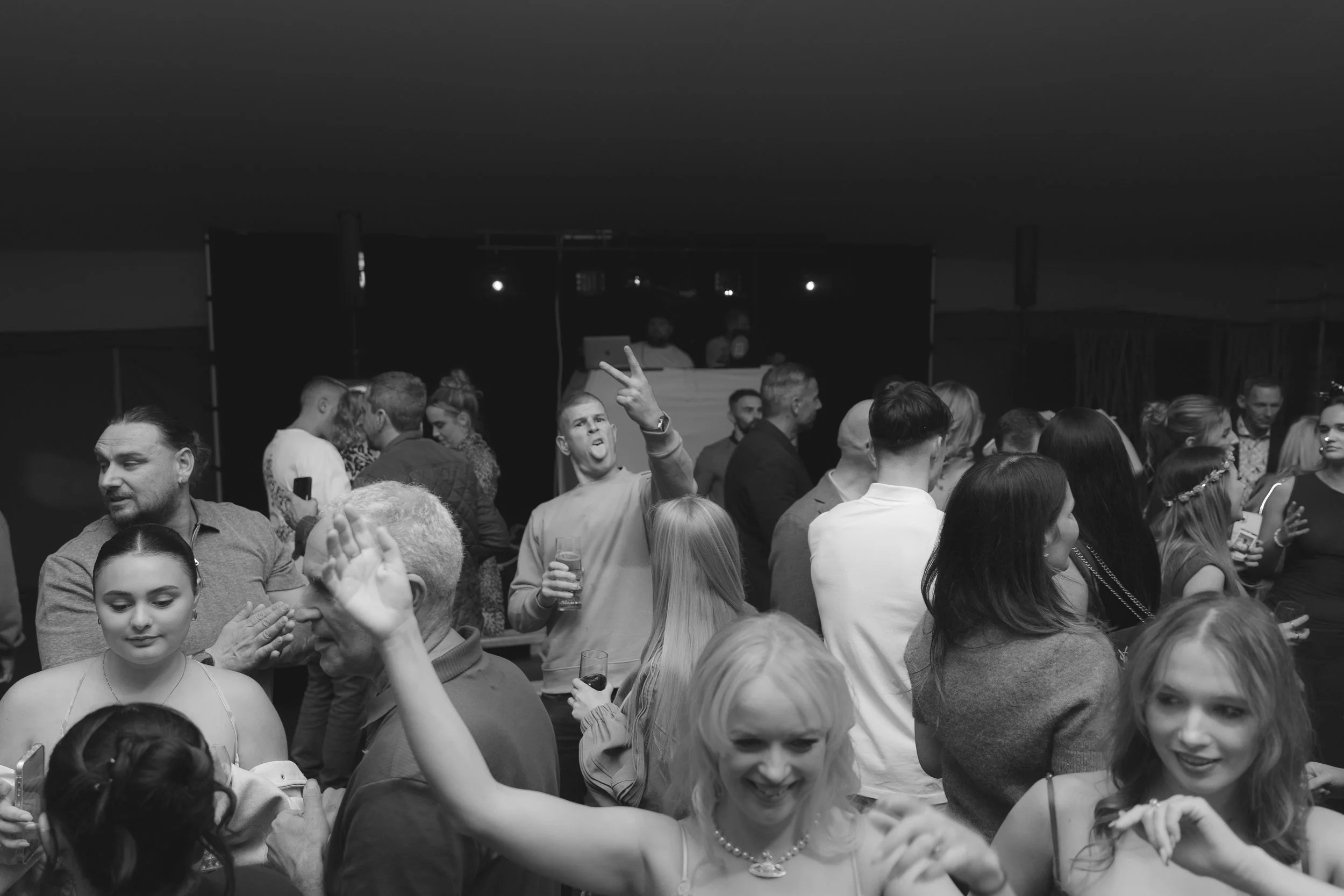 Crowd of people dancing and socializing at a party with DJ in the background, black and white photo.