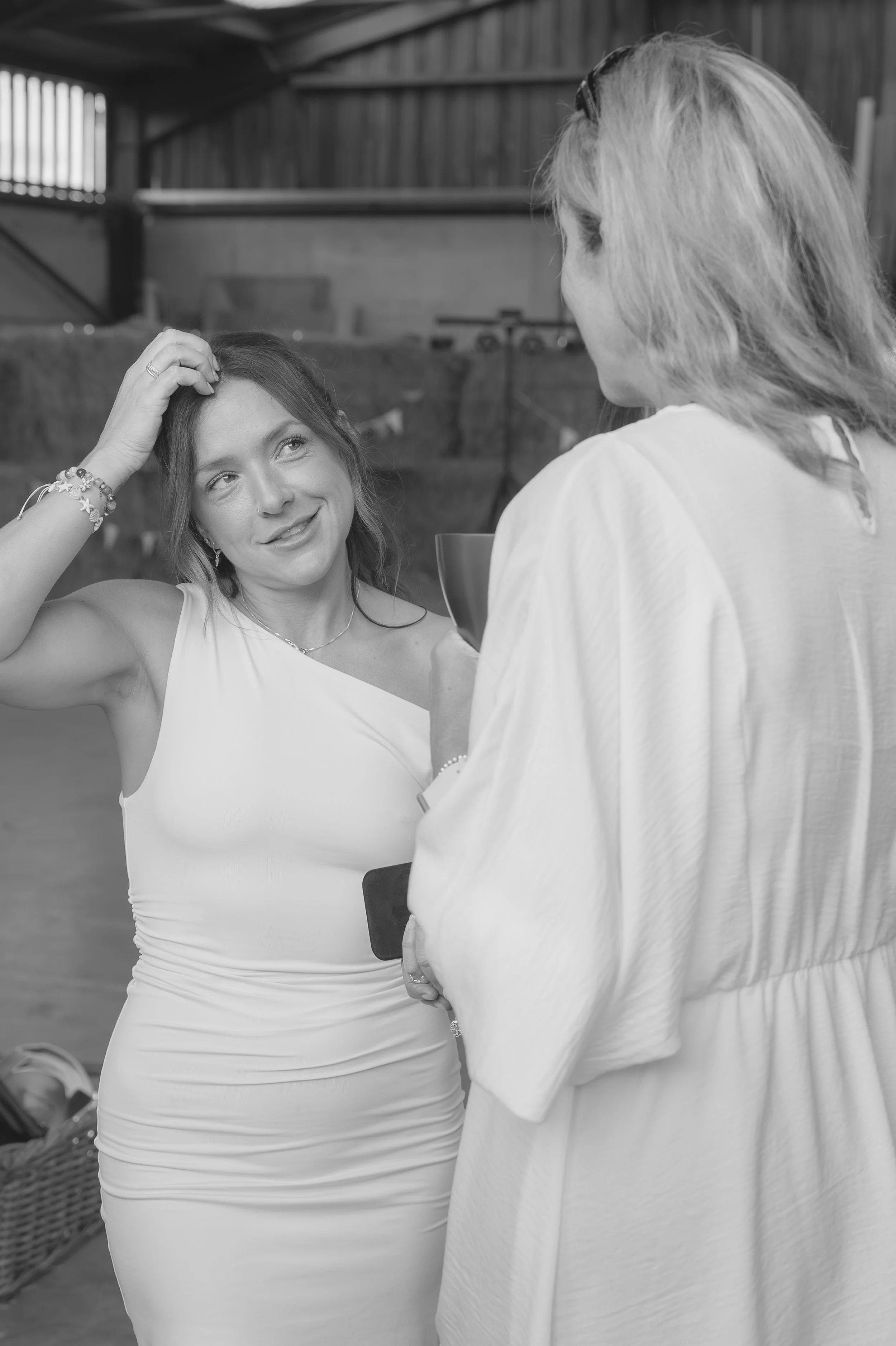 Two women engaged in conversation in a barn-like setting. The woman on the left is smiling, with her hand on her head, wearing a sleeveless dress and bracelets. The woman on the right, holding a drink, has long hair and is wearing a light-colored top