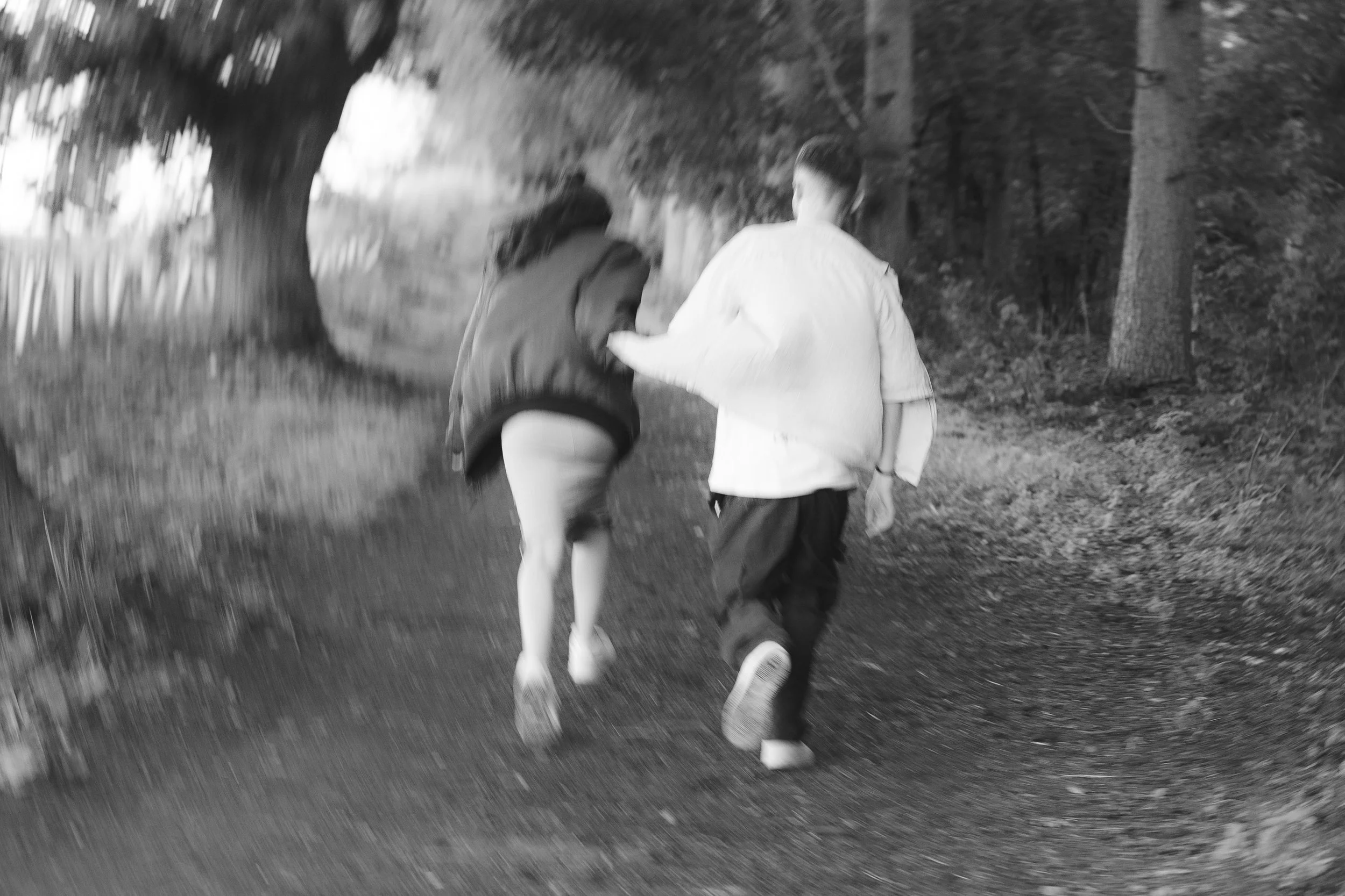 Two children walking together along a sidewalk in a park, one wearing a jacket and shorts, the other in a long-sleeve shirt and pants, near trees and greenery.