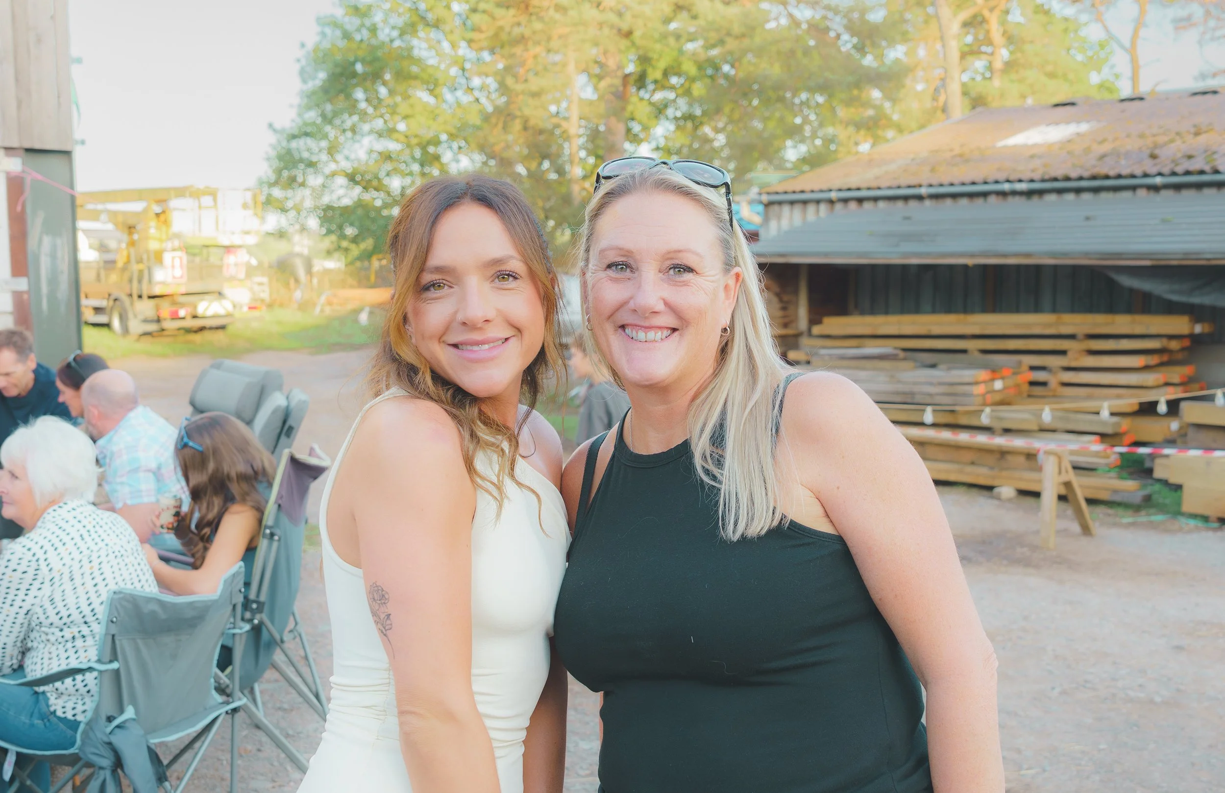 Two women smiling outdoors at a gathering, with trees and a shed in the background.