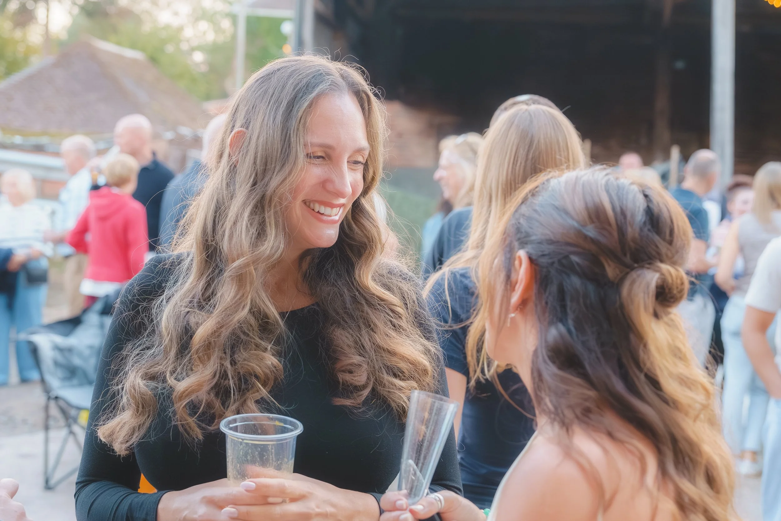 A woman with long, curly blonde hair smiling and holding a plastic cup, engaged in conversation with another woman at an outdoor event surrounded by other people.