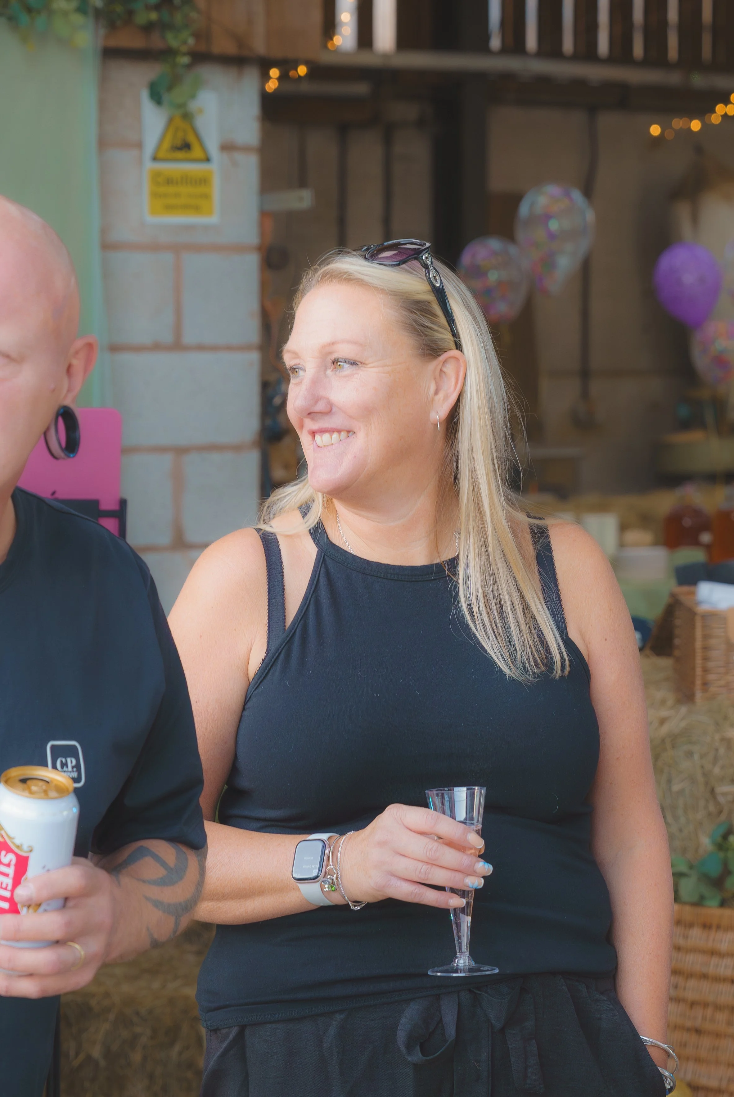 A woman with blonde hair, sunglasses on her head, wearing a black sleeveless top, is smiling and holding a champagne flute. She wears a silver smartwatch and jewelry. Part of a man with tattoos, earrings, and a black shirt is visible. The background 