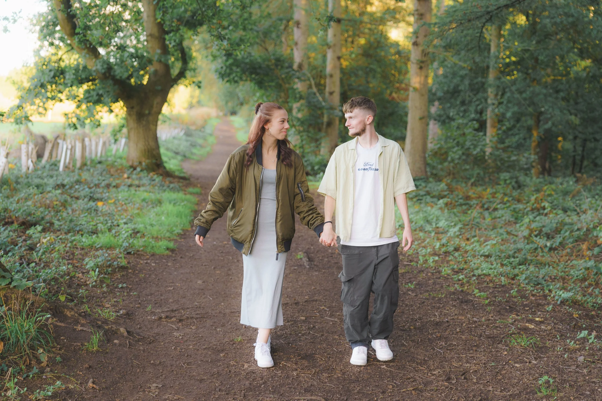 A young couple walking hand in hand on a forest trail surrounded by tall green trees and lush foliage.