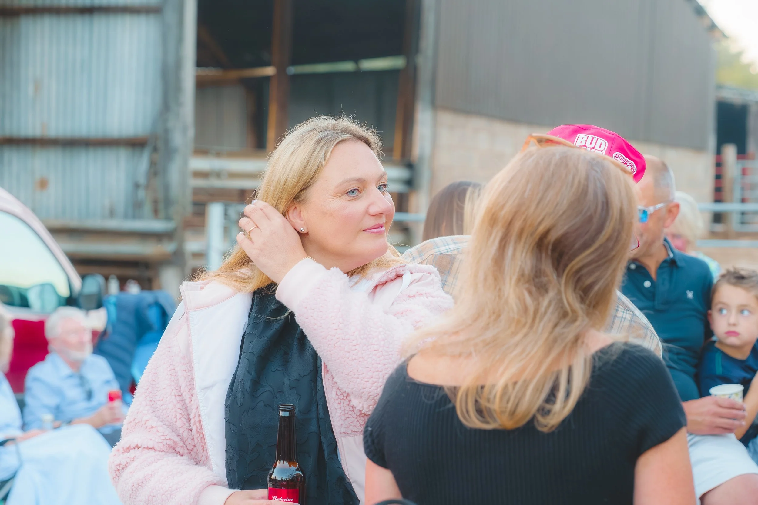 A woman with blonde hair and a pink jacket, holding a bottle of Budweiser beer at an outdoor gathering with other people in the background.