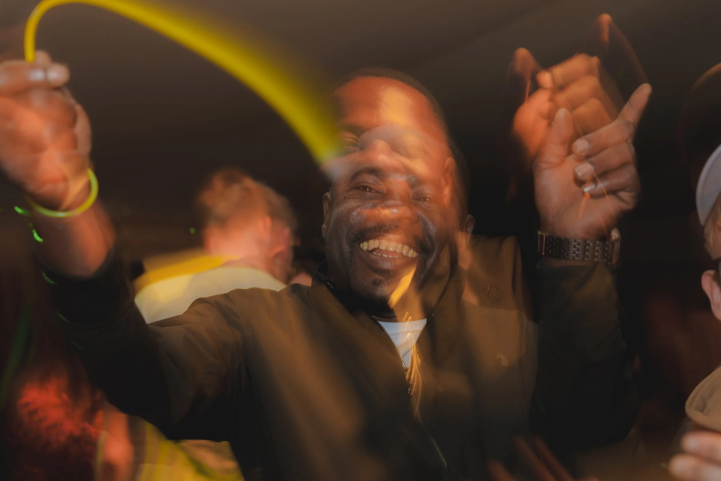 A smiling man with dark hair taking a selfie with a group of people, all appearing joyful, in a lively setting with warm lighting and some motion blur.