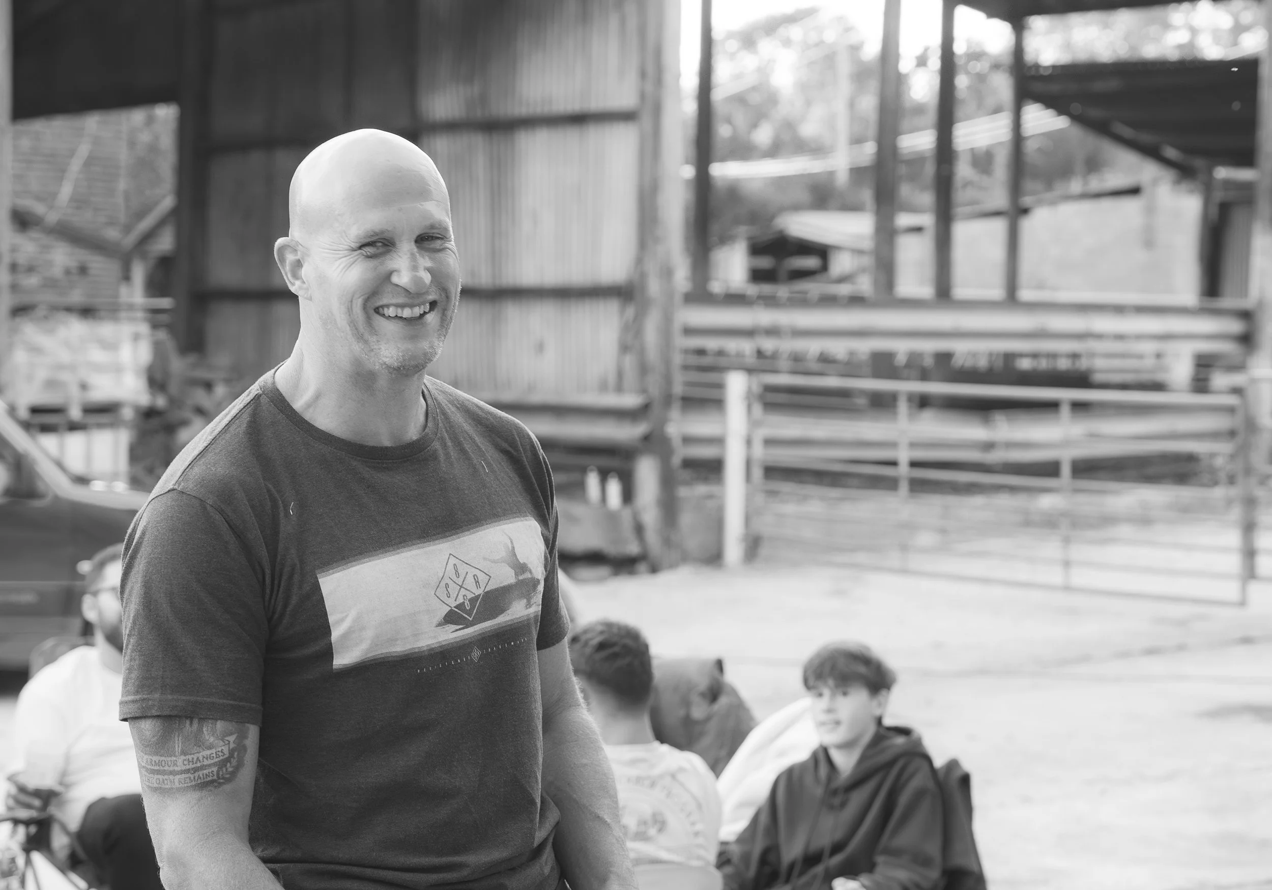 Black and white photo of a middle-aged man with a bald head smiling and winking at the camera. He is wearing a T-shirt with a graphic design. There are children sitting behind him, and structures like bleachers and a building are visible in the backg