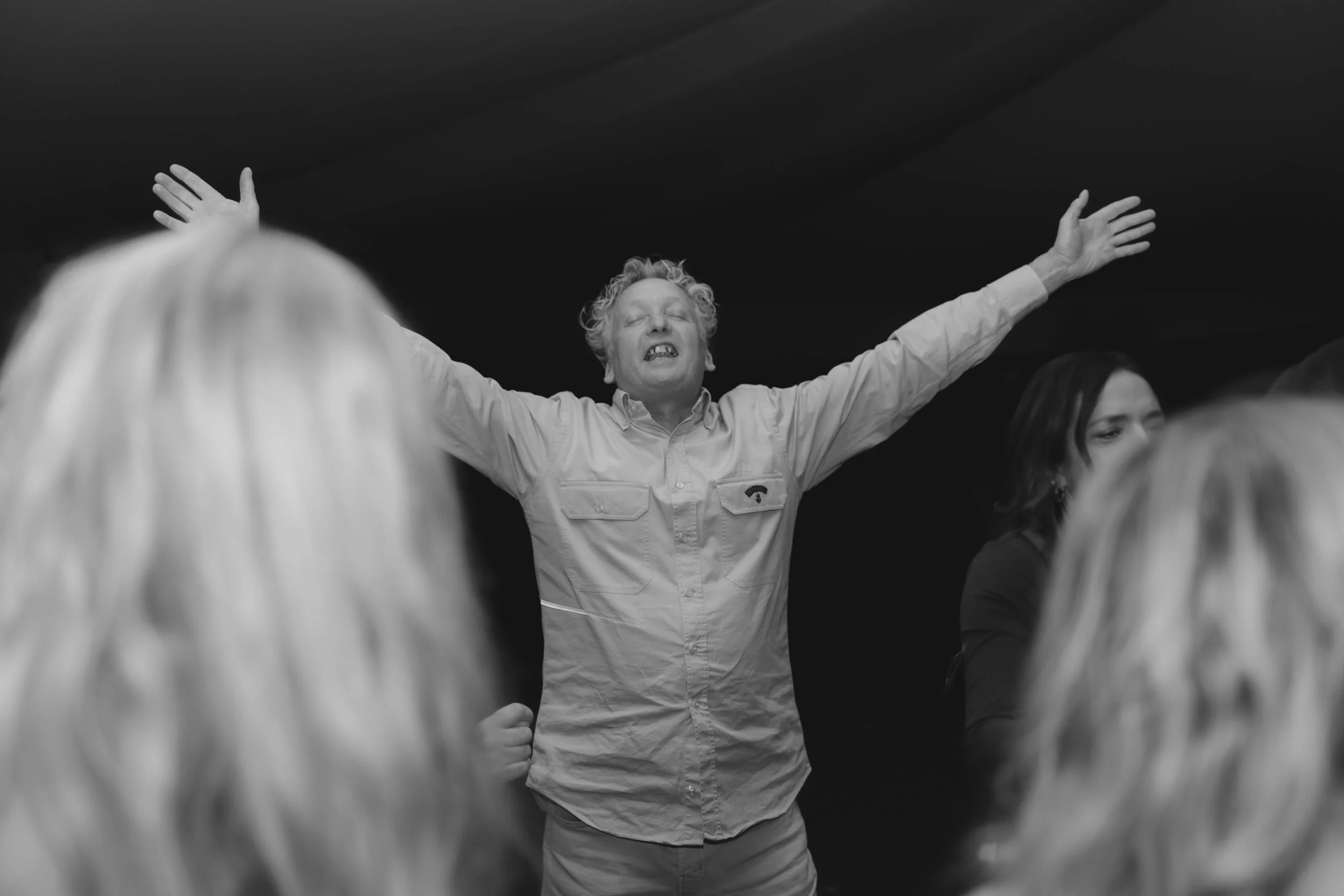 A man with curly hair and outstretched arms appears to be leading or speaking to a group, with people in the foreground and a dark background.