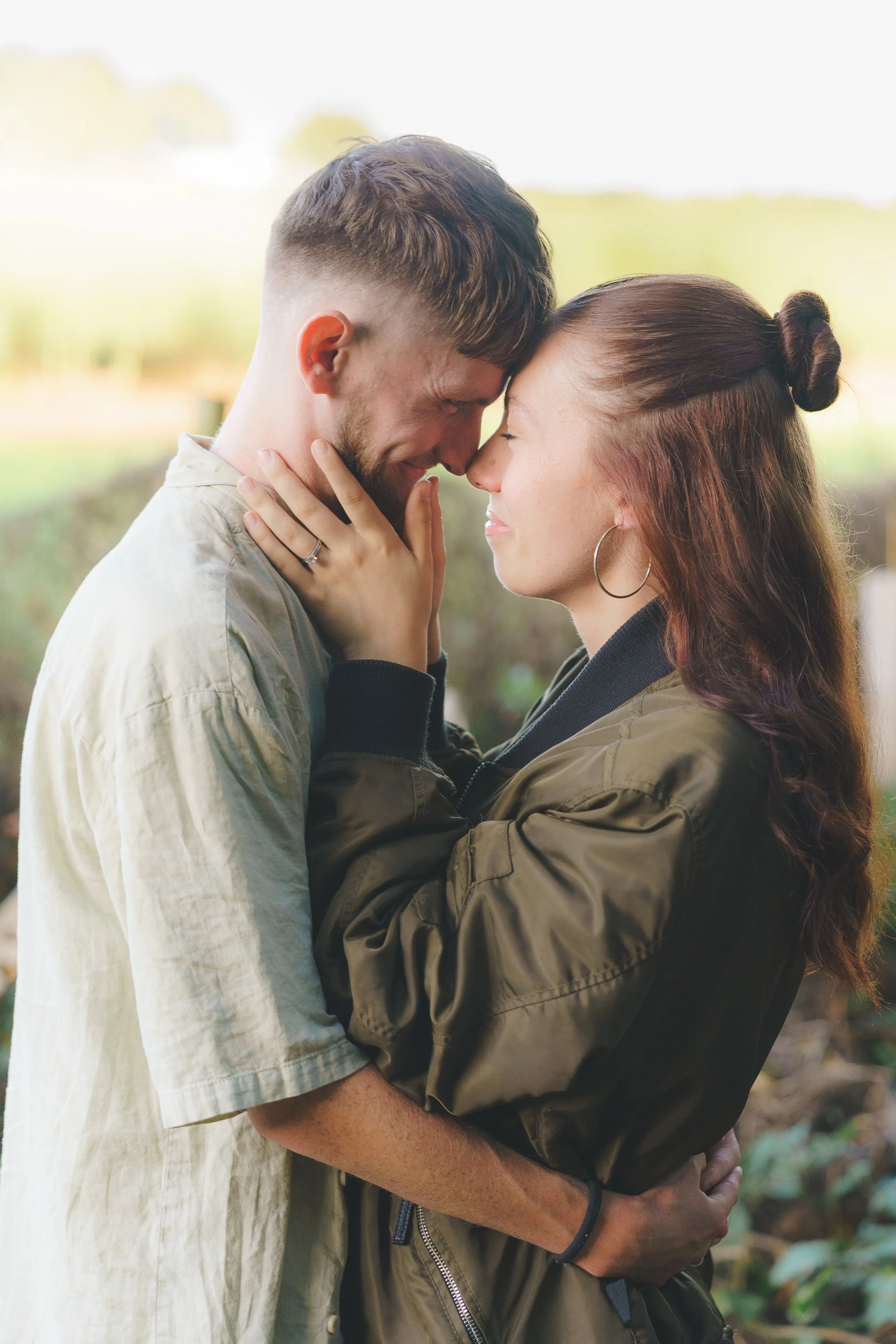 A romantic couple with their foreheads touching, holding each other outdoors in a park-like setting.