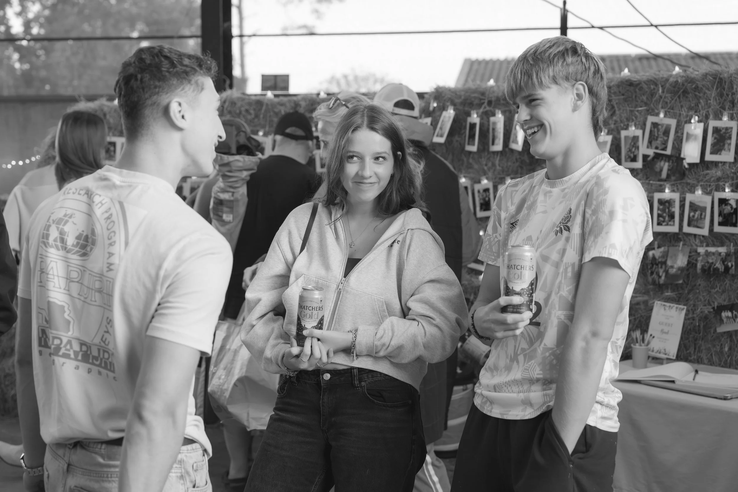 Three teenagers, two boys and a girl, are talking and smiling at each other at an outdoor event. The girl is wearing a hoodie and holding a can of drink, while the boys are in casual T-shirts. There are photos displayed on a string background behind 