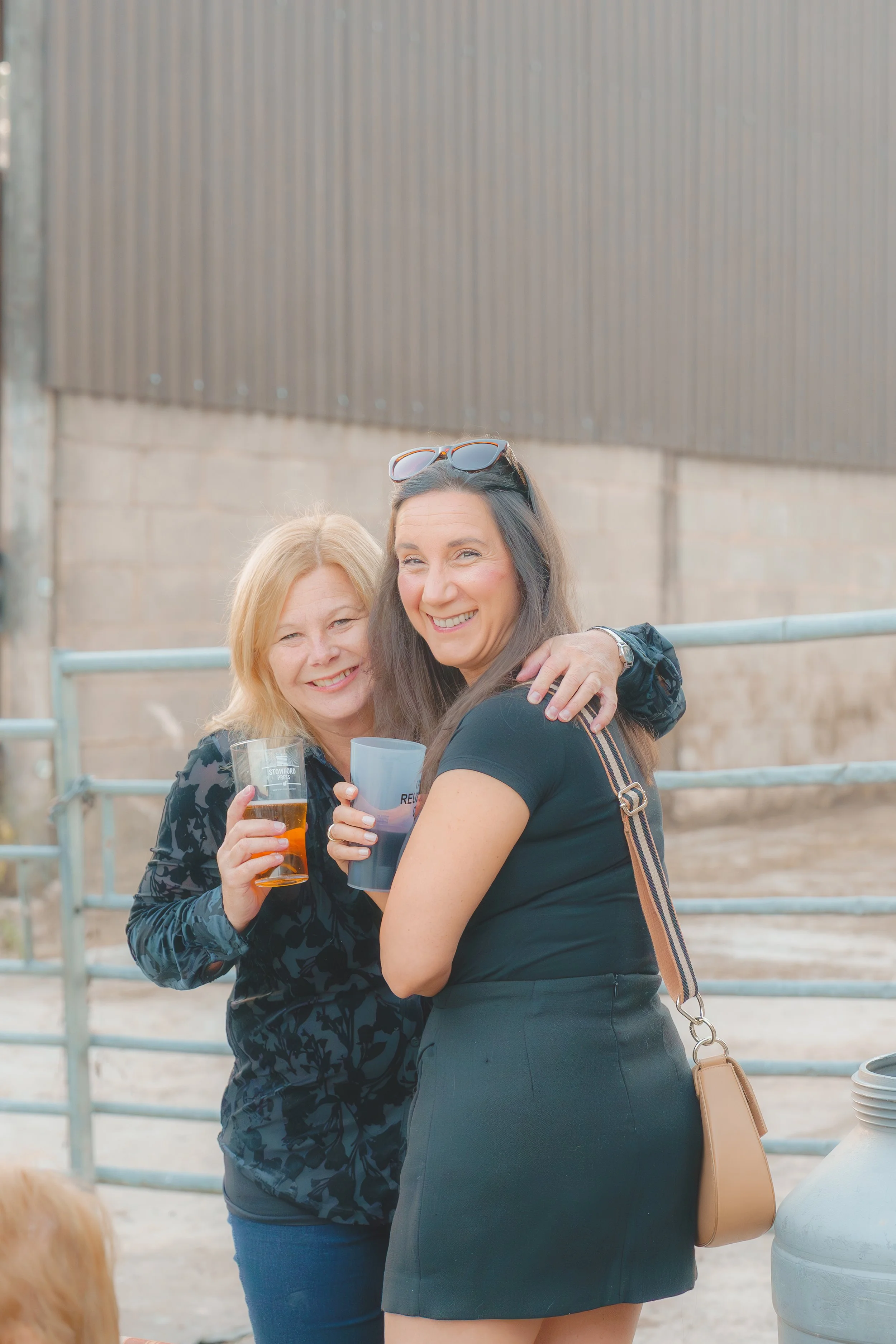 Two women smiling and hugging each other outdoors, holding drinks, standing near a metal fence and a large container.