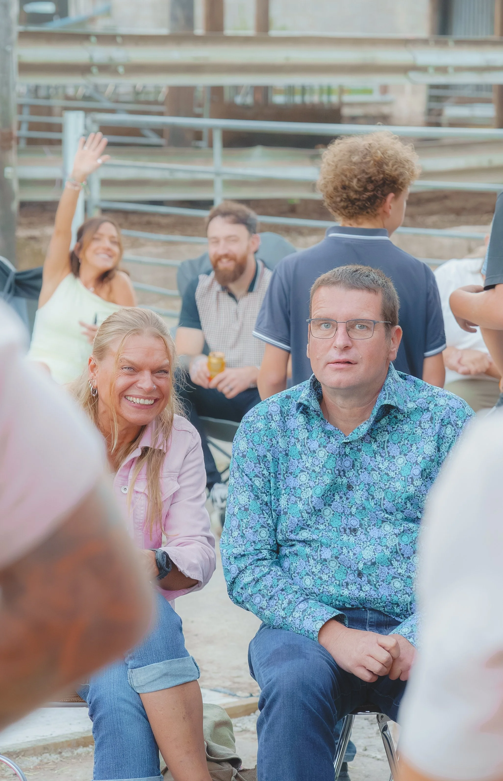 A group of people gathered outdoors, sitting and chatting, with a woman waving in the background. The setting appears casual and social.