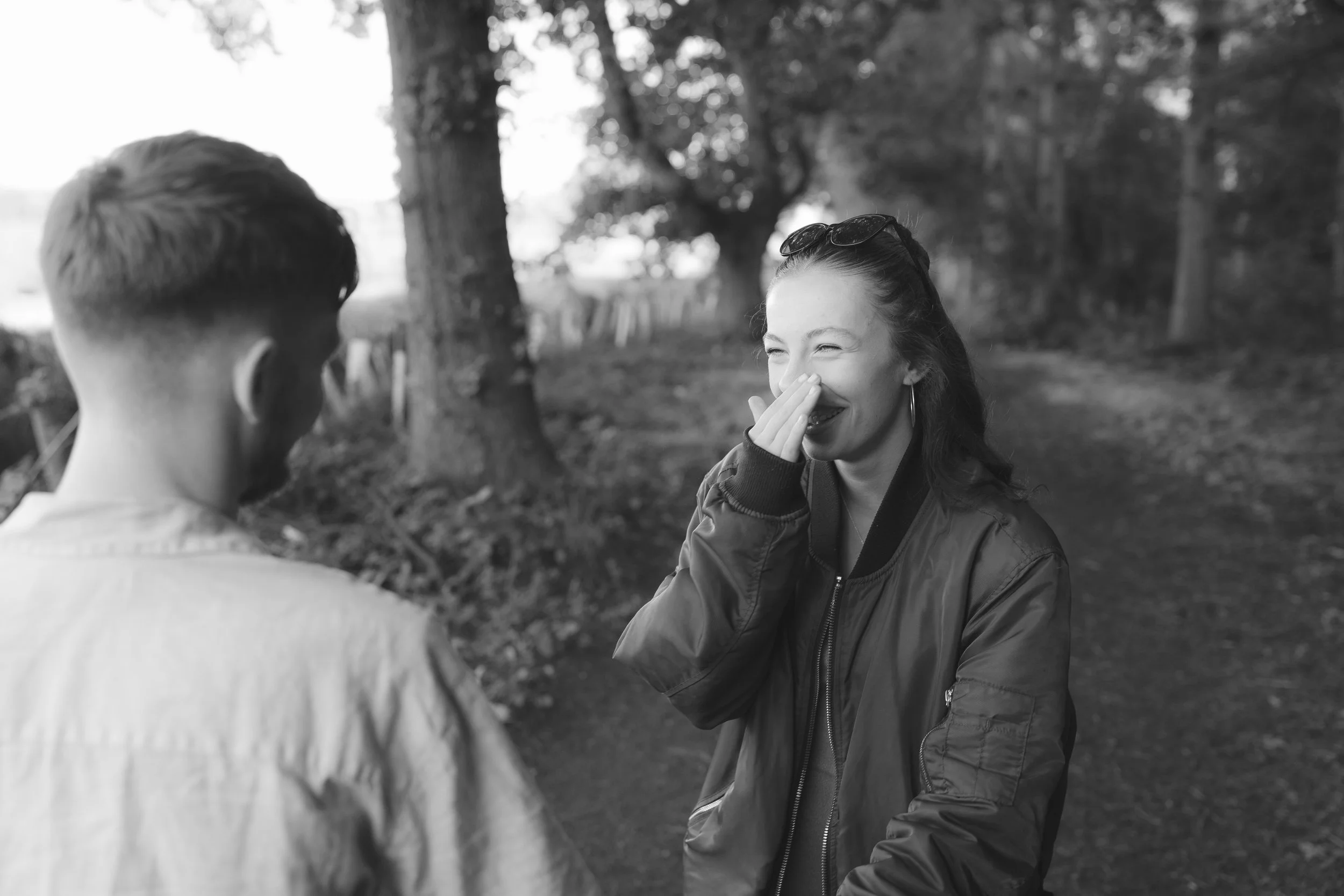 A woman with glasses on her head laughs and covers her mouth with her hand while talking to a person with short hair outdoors in a park with trees and a fence in the background, in black and white.