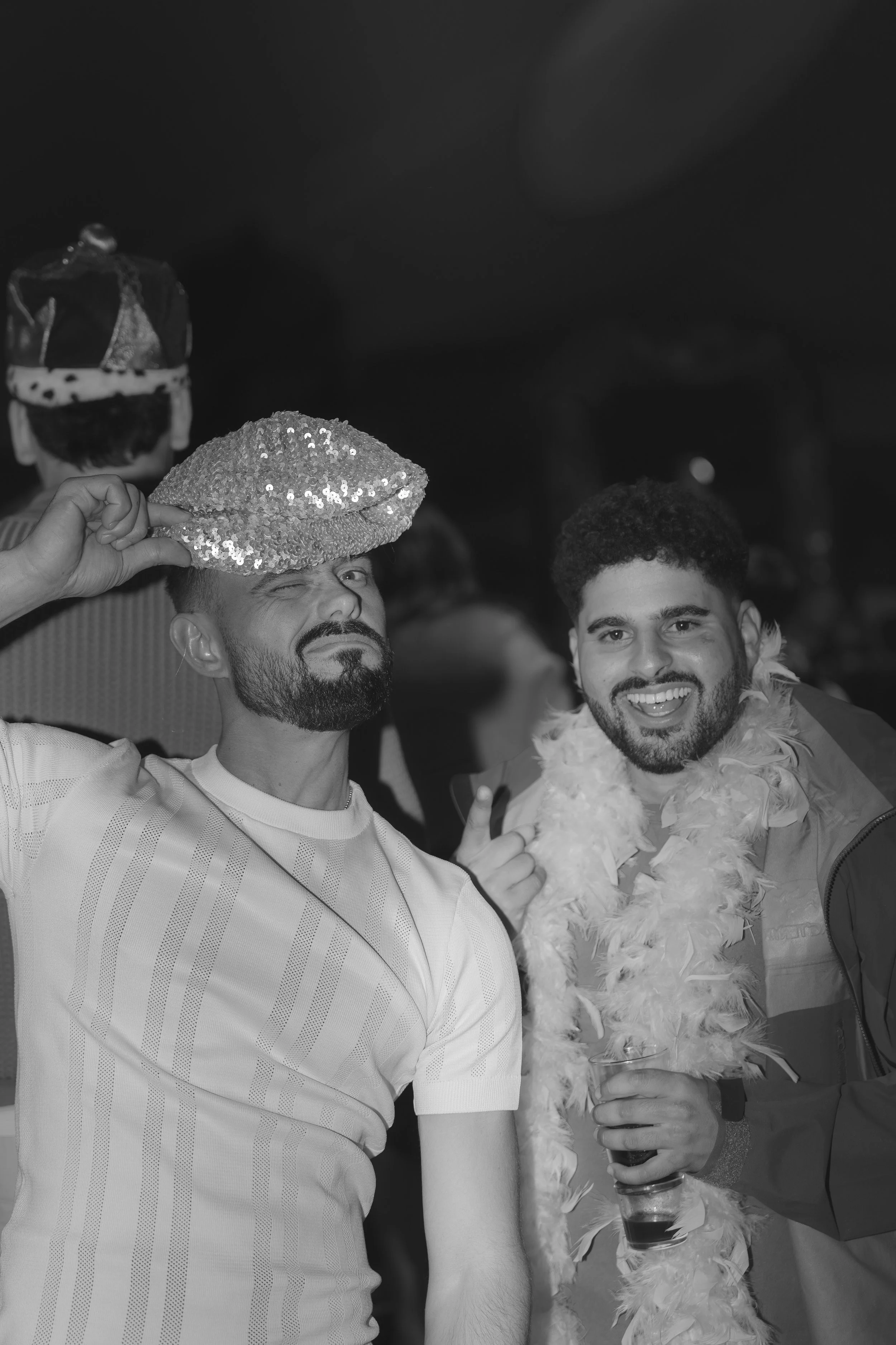 Two men at a party, one wearing a shiny sequin hat and the other with a feather boa, holding drinks and smiling.