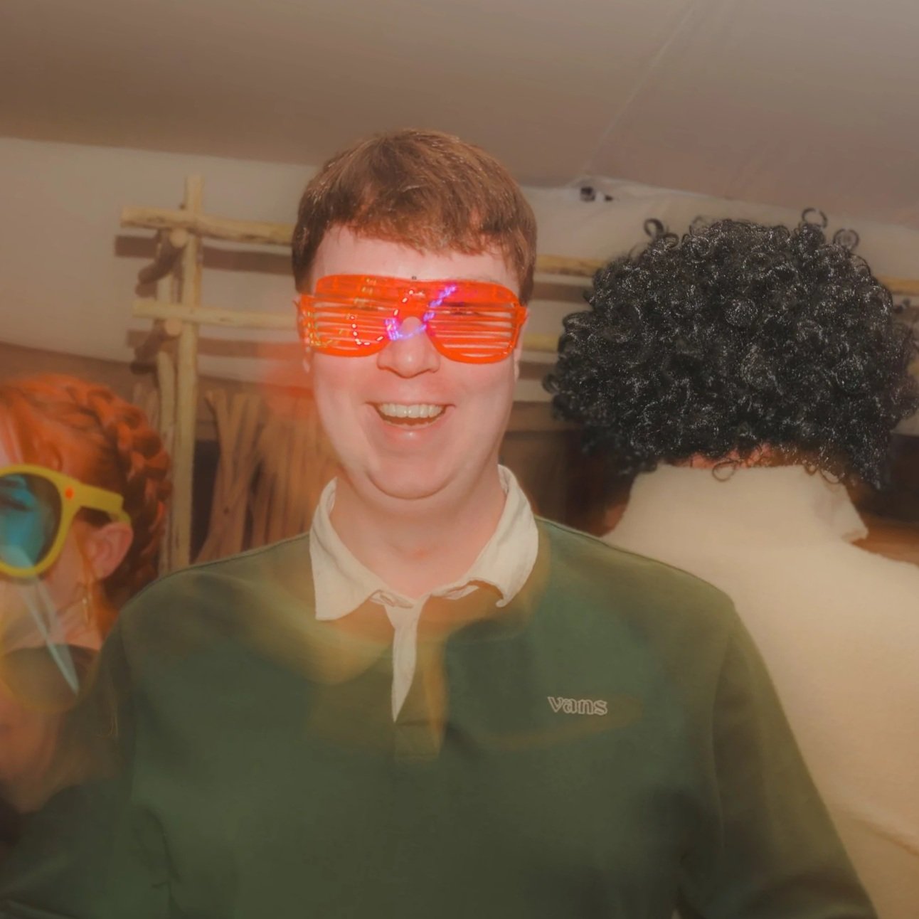 A man smiling wearing orange shutter shades, a white collared shirt under a green Vans sweatshirt, in a room with people wearing colorful wigs and sunglasses in the background.