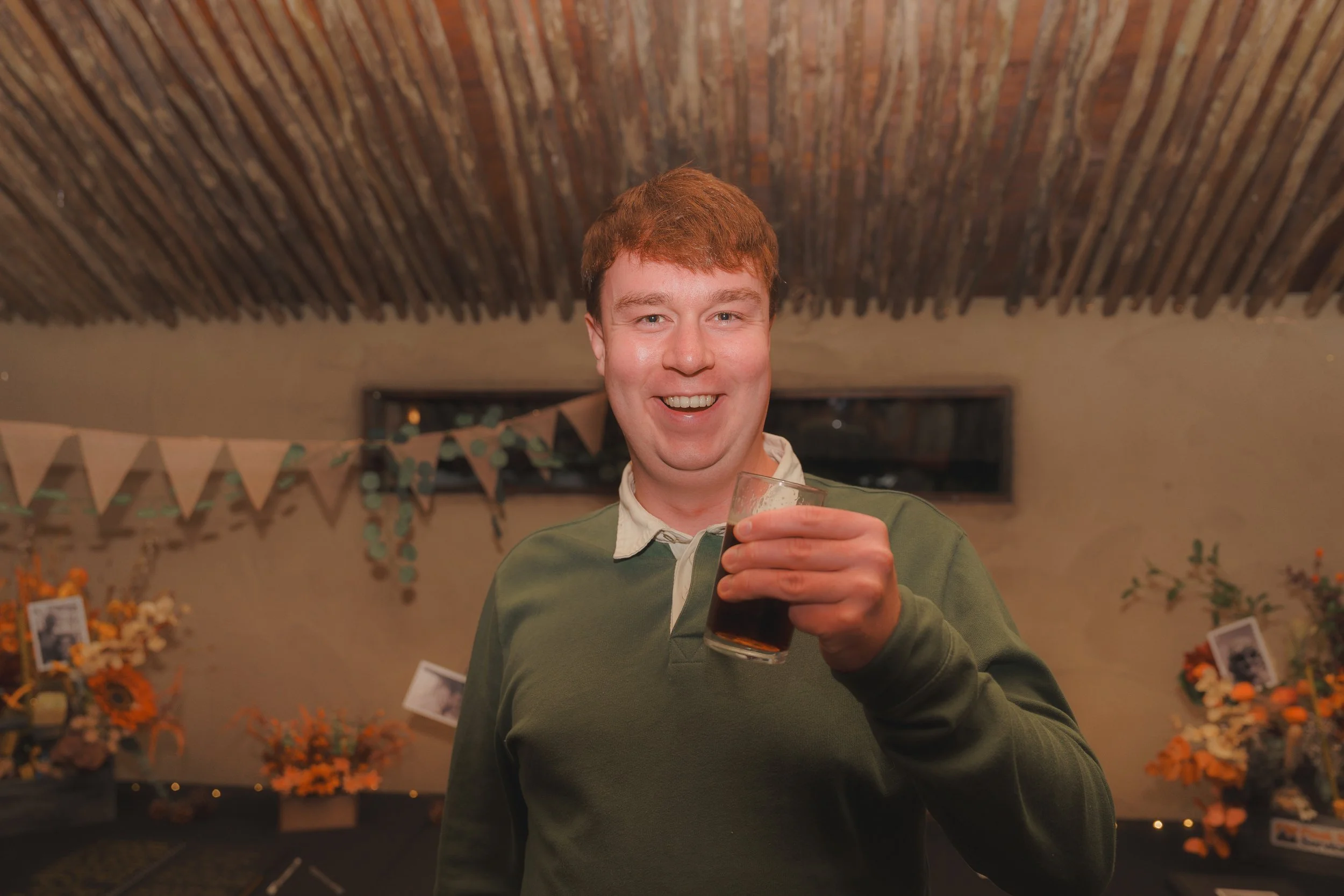 A young man with red hair smiling and holding a glass of dark beer in a decorated indoor setting.