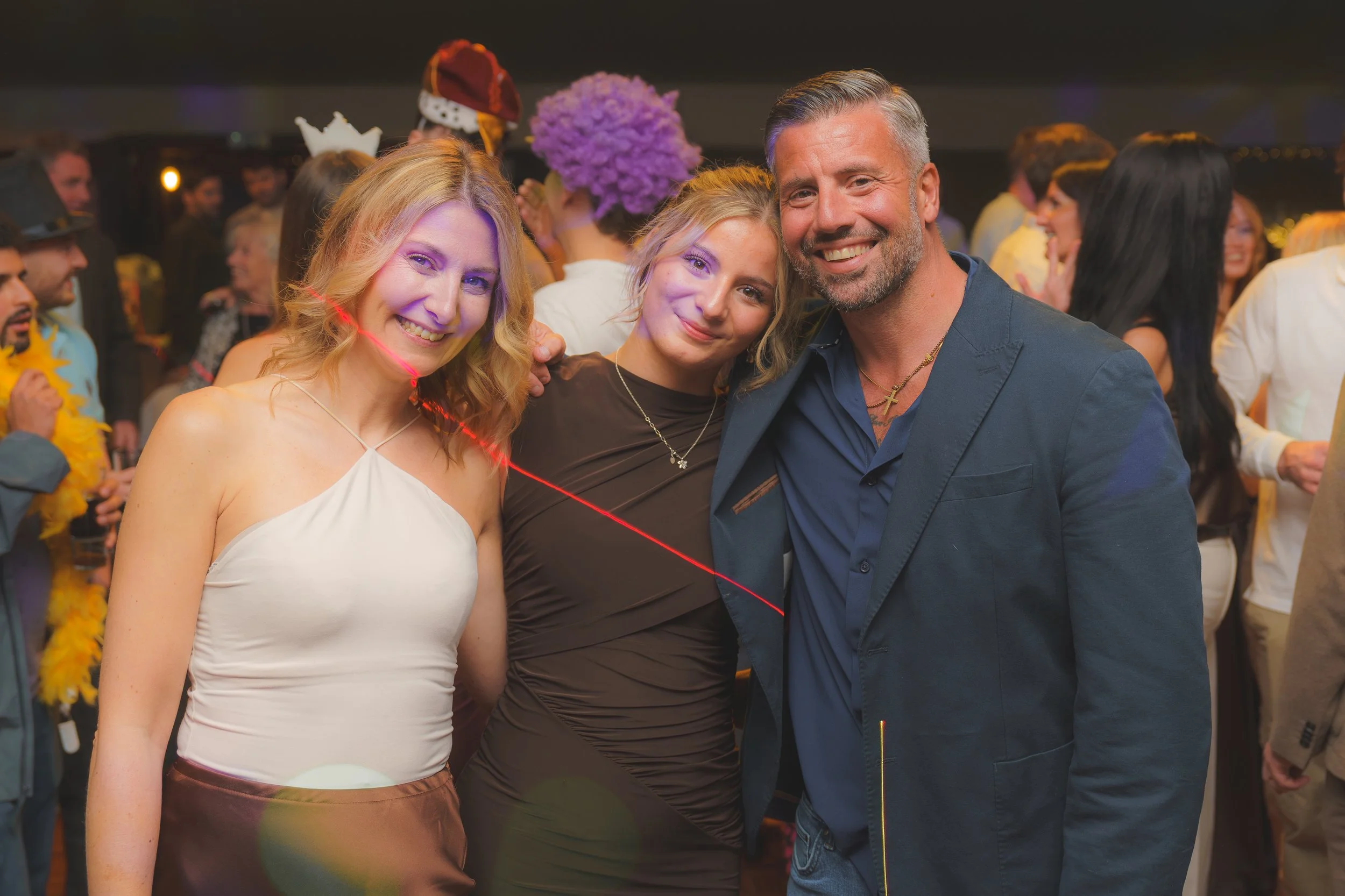 Three people smiling at a party with colorful costumes and decorations in the background.