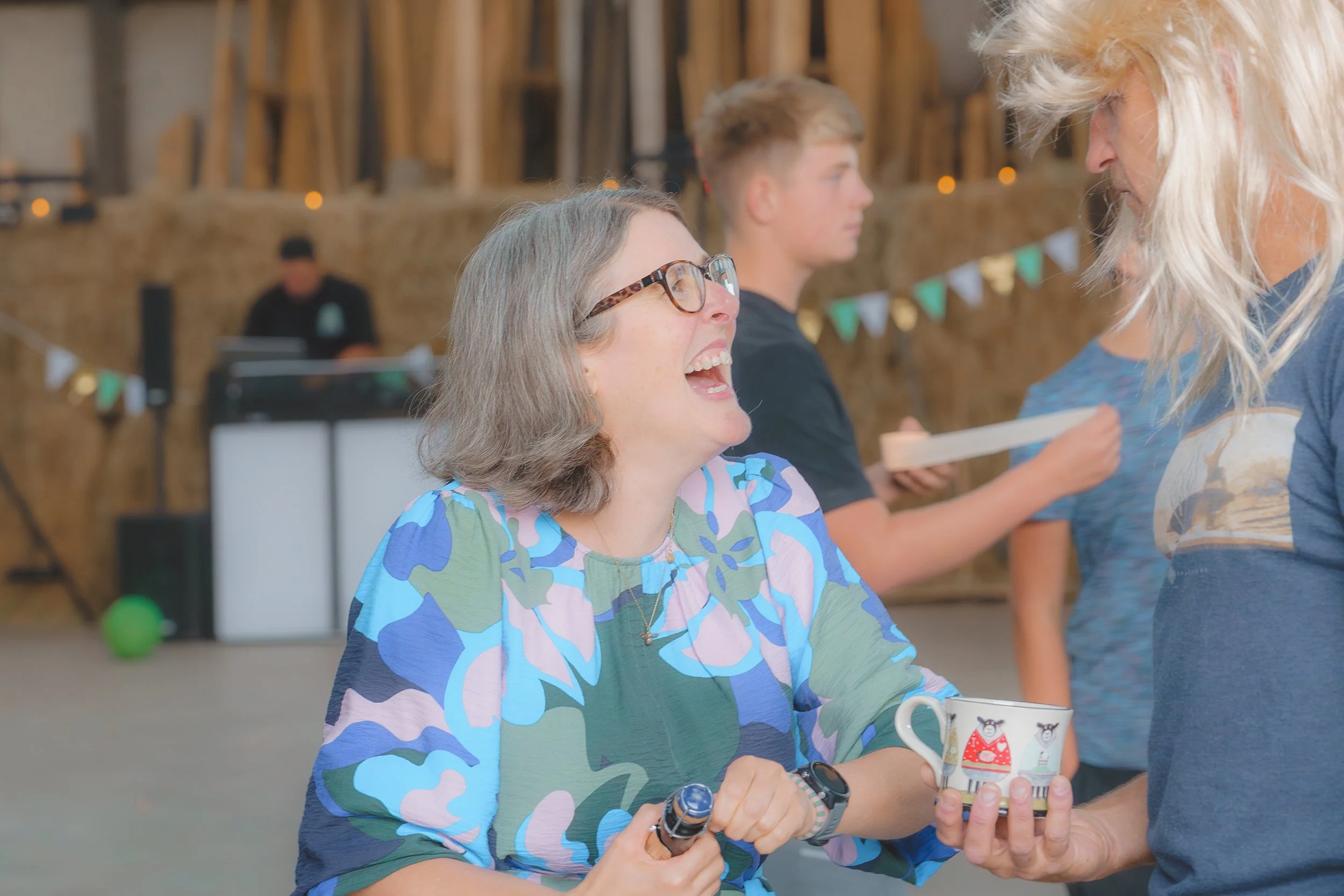A woman with gray hair and glasses laughing and talking with a blonde woman at a gathering, with a young man standing in the background, in a rustic indoor setting decorated with bunting and string lights.