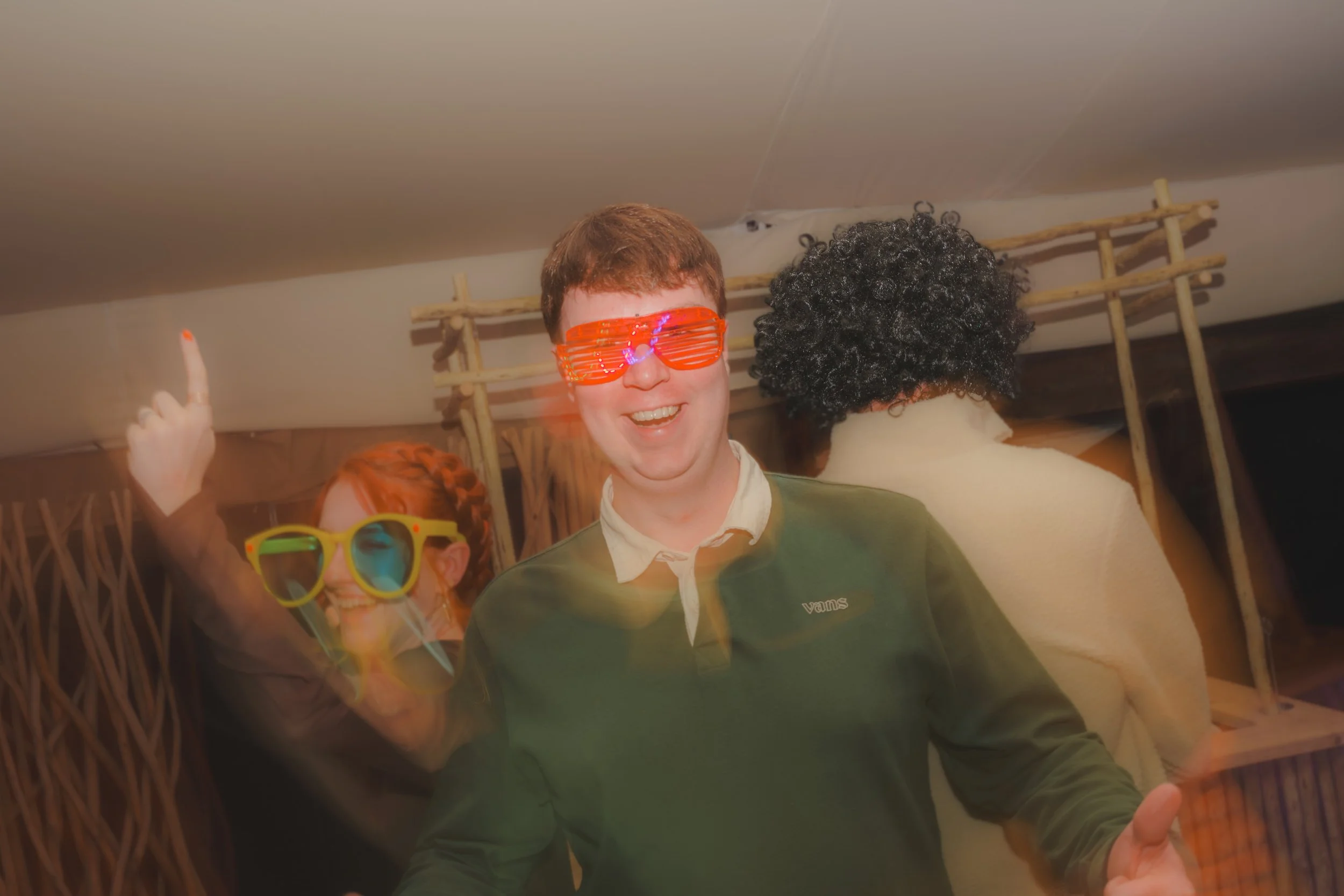 Two people celebrating at a party, wearing colorful sunglasses, one with red glasses and the other with yellow glasses, smiling and having a good time.