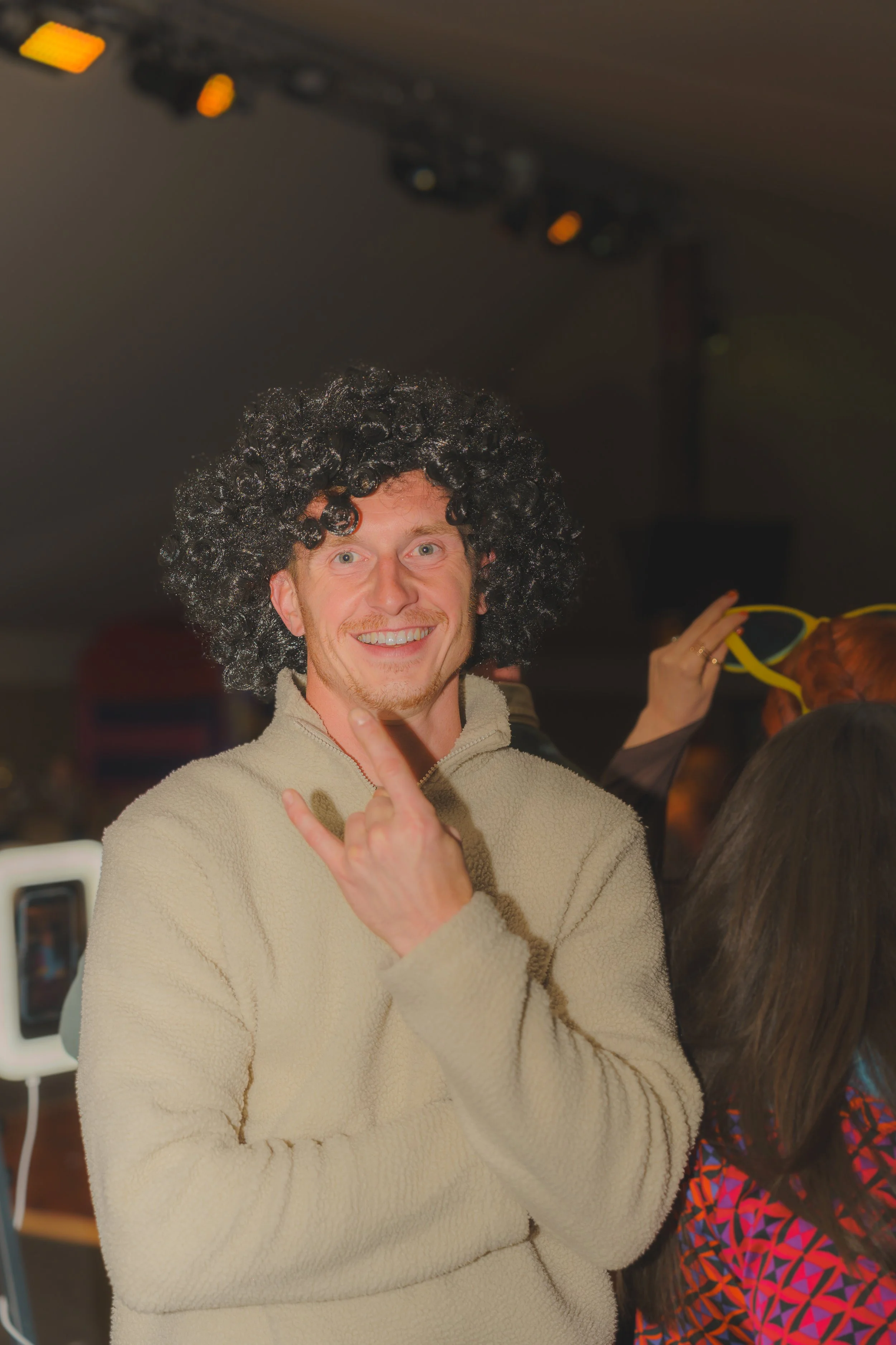 A young man with a curly black wig smiling and giving a peace sign in an indoor setting. He is wearing a beige fleece jacket, and there's a woman in the background with long brown hair in colorful patterned clothing.