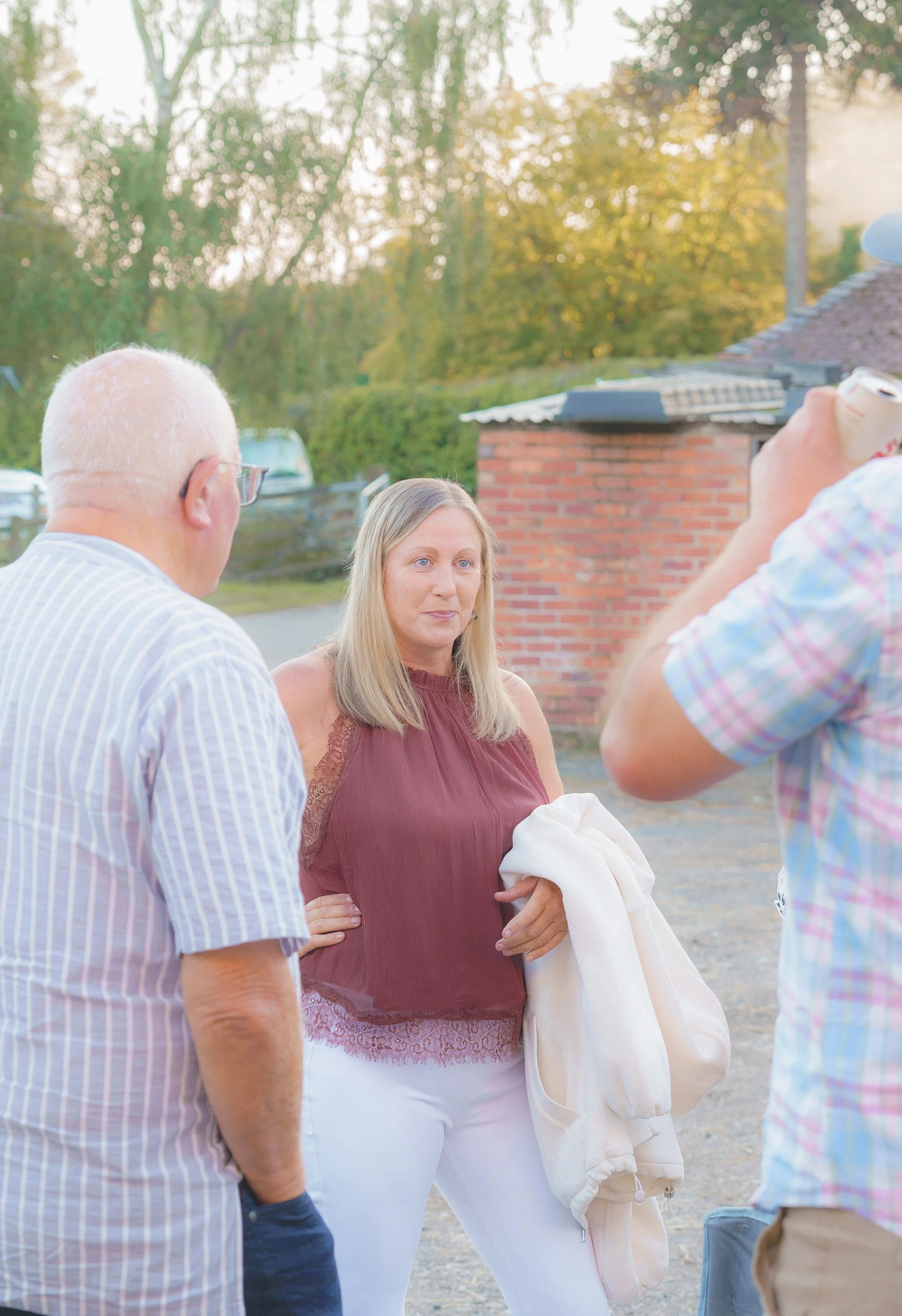 A group of people engaging in conversation outdoors during the daytime, with trees and a brick building in the background.